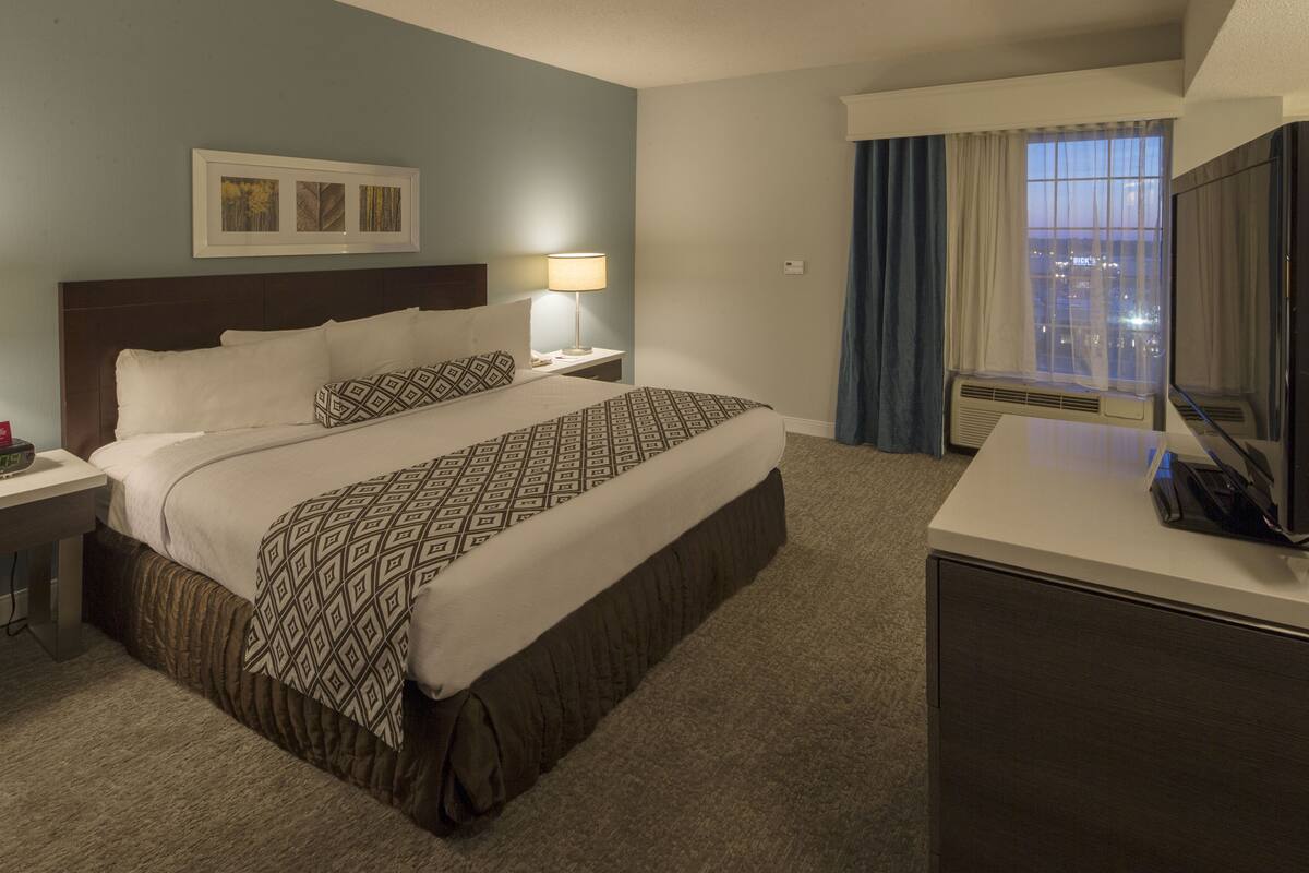 Ein Hotelzimmer mit Bett, Nachttisch, Lampe, Fernseher und einem Fenster mit Vorhängen.