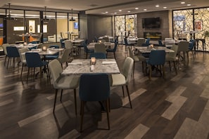 A modern dining area with multiple tables and chairs, a flat-screen TV mounted on the wall, and a decorative wall with geometric patterns.