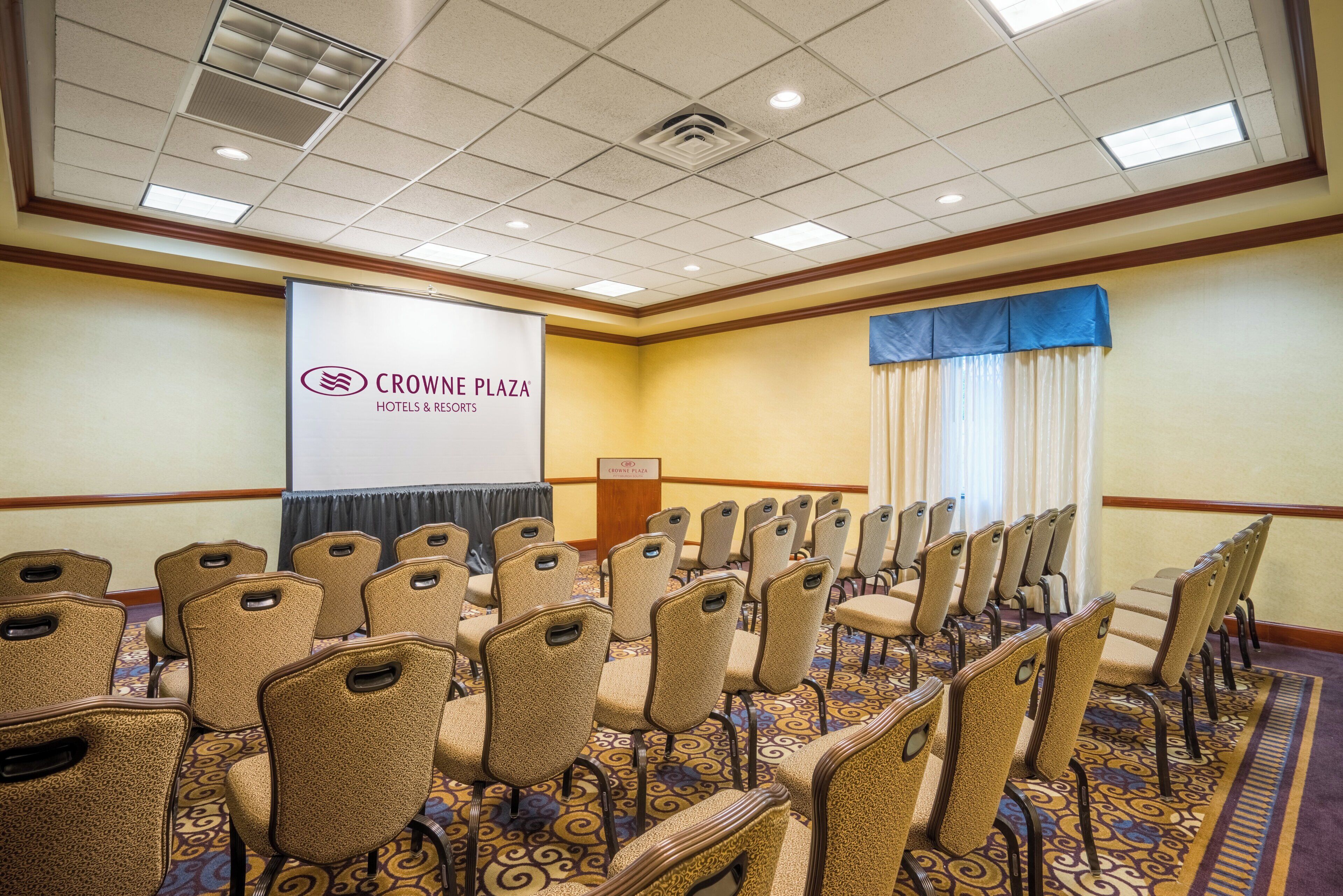 Un salón de conferencias con filas de sillas, un atril y un cartel de Crown Plaza Hotels & Resorts.