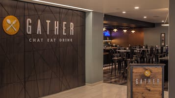 Et moderne interiør med et skilt der står 'GATHER CHAT EAT DRINK' på væggen, et spiseområde med borde og stole, samt et træskilt med samme navn.