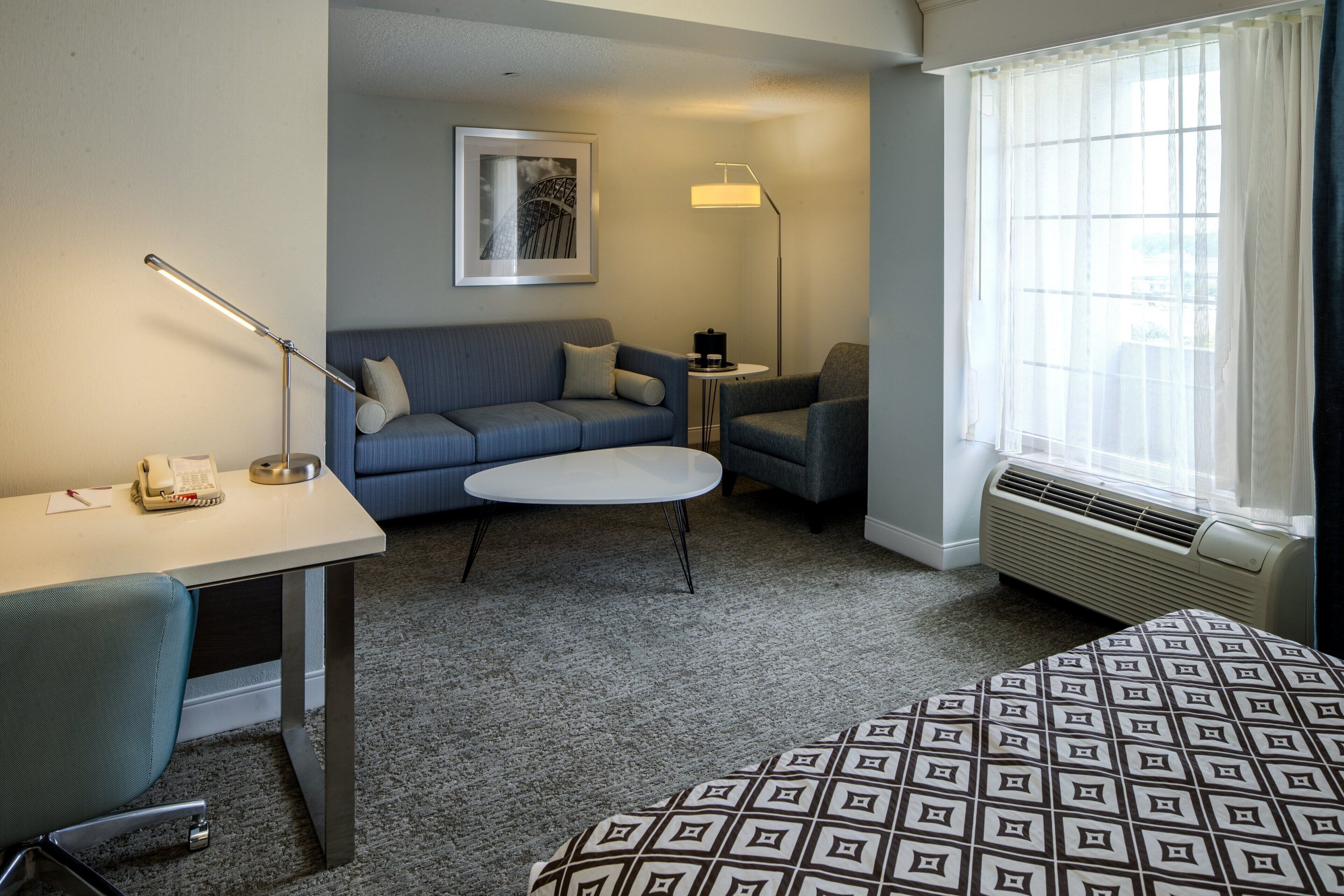 A hotel room with a bed, a desk with a phone, a couch, a small round table, and a window with curtains.