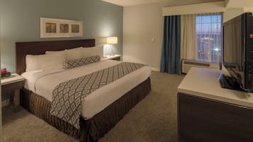 A hotel room with a bed, a desk with a television, a nightstand with a lamp, and a window with curtains.