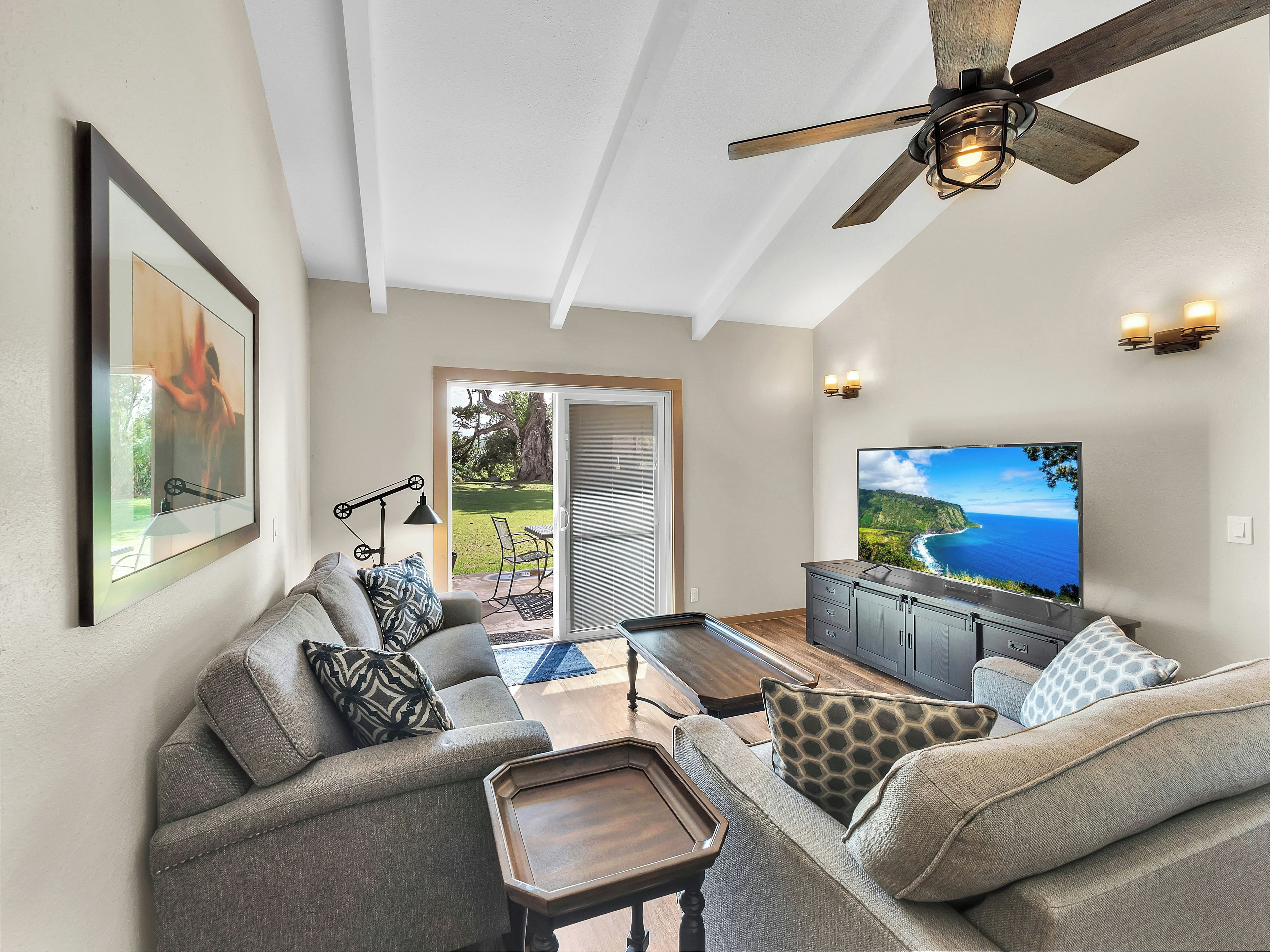 A modern living room with a ceiling fan, a large flat-screen TV, and a view of the outdoors.