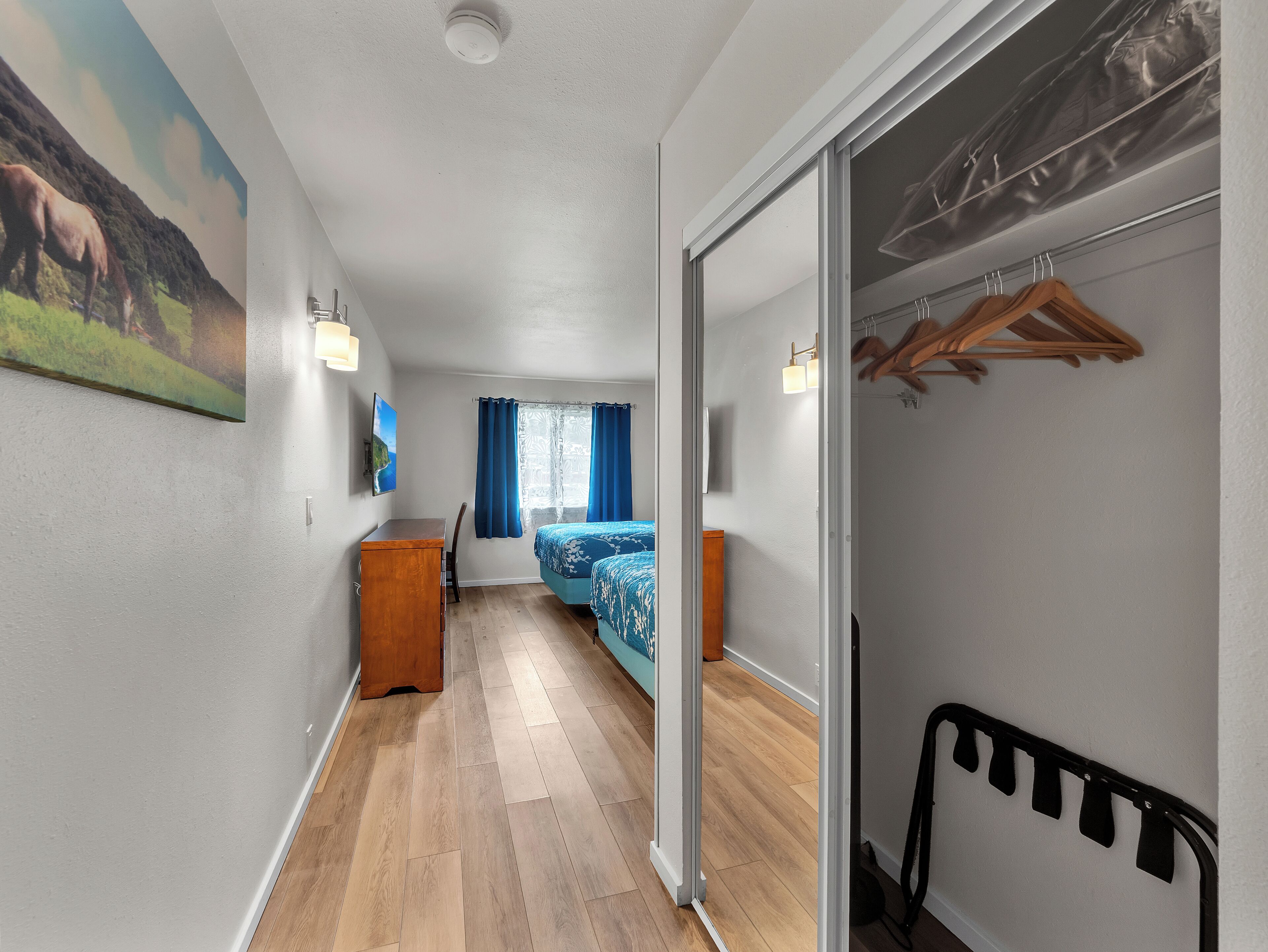 A narrow hallway with a wooden desk, a bed with a blue bedspread, and a wardrobe with wooden hangers.