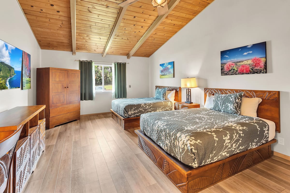 A bedroom with two beds, wooden ceiling beams, a ceiling fan, a window with curtains, and artwork on the walls.