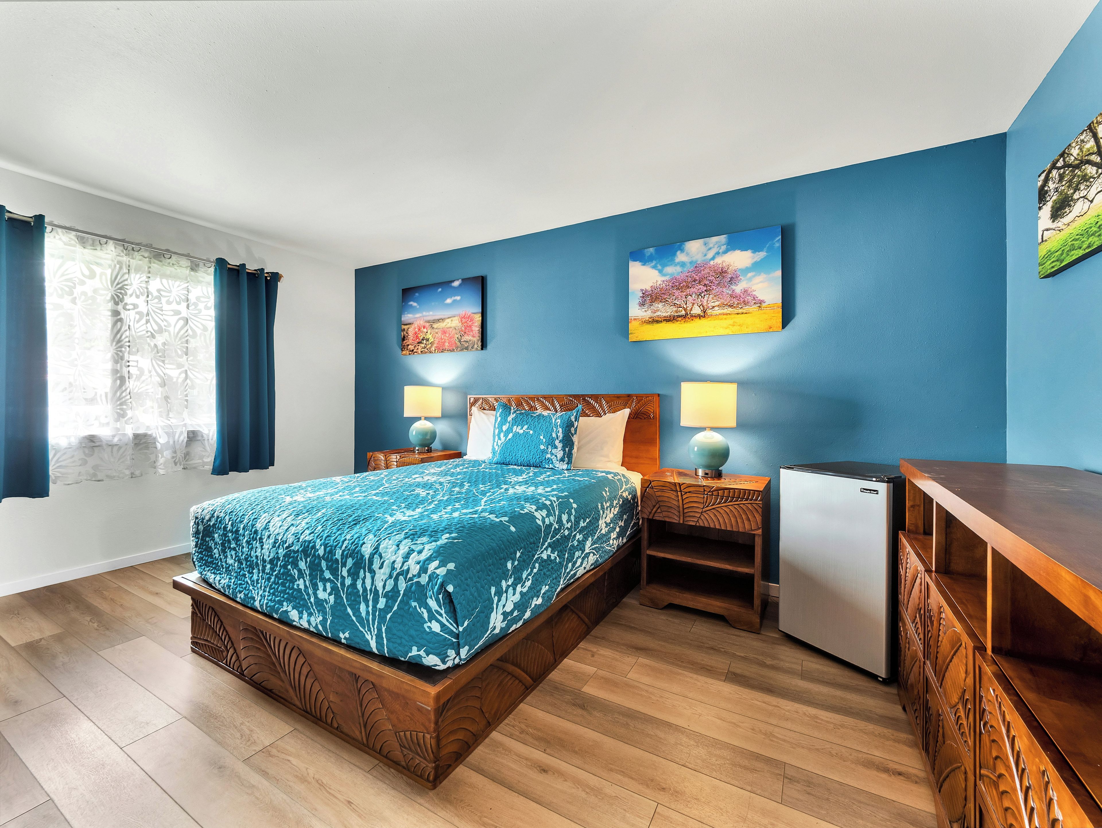 Un dormitorio con una cama, dos mesitas de noche, una lámpara, un refrigerador pequeño y un aparador de madera.
