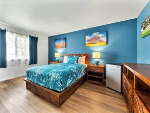 A bedroom with a bed, two bedside tables, a lamp, a small refrigerator, and a wooden dresser. - Castle Waimea Country Lodge (Kamuela)