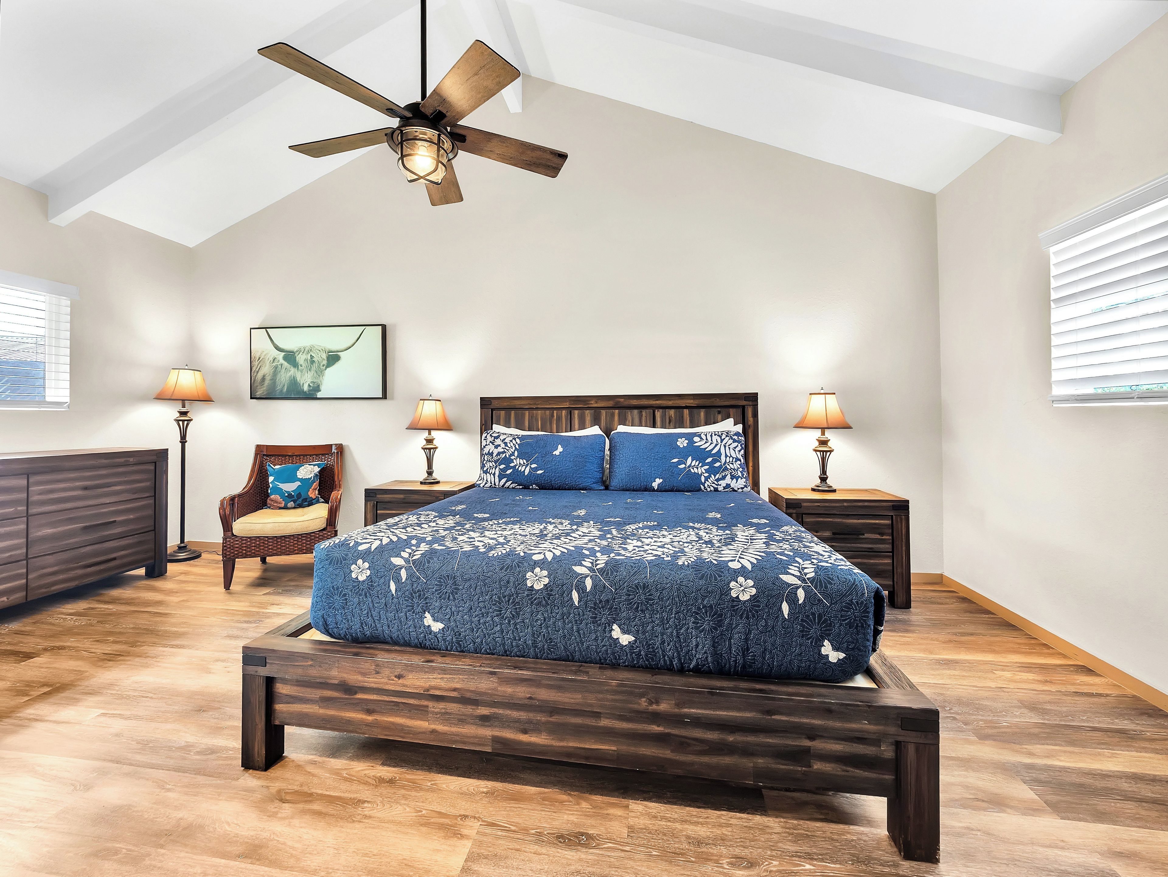 Un dormitorio con cama de madera, dos lámparas de noche, ventilador de techo y un ventanal con persianas.
