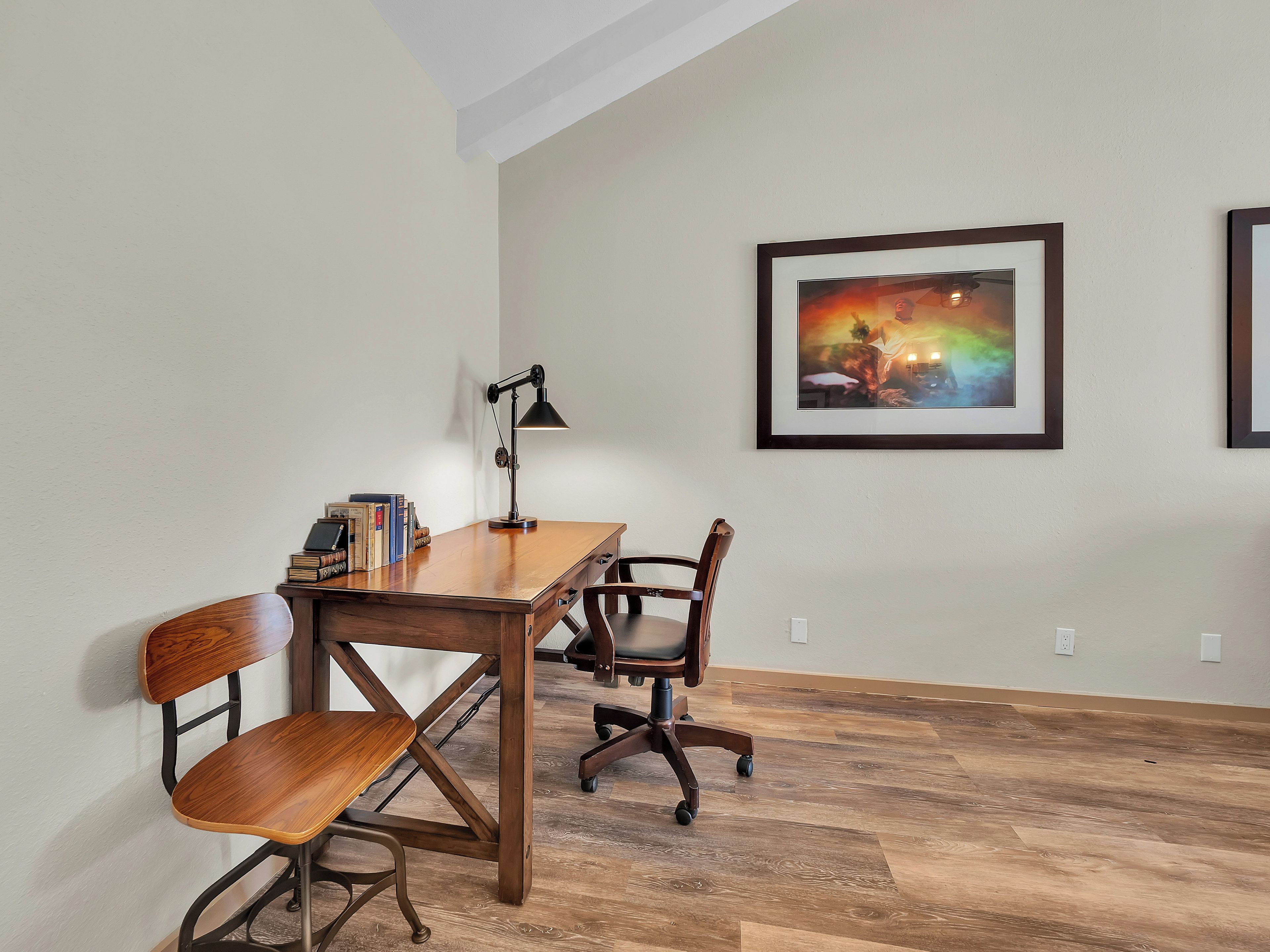 A room with a wooden desk, a chair, a lamp, and a framed picture on the wall.