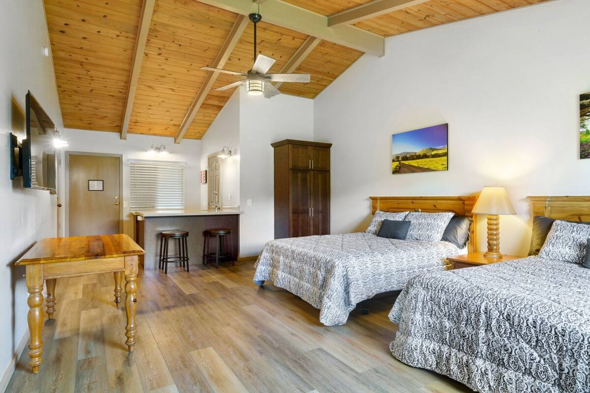 A room with two beds, a wooden ceiling, a desk with stools, and a painting on the wall.
