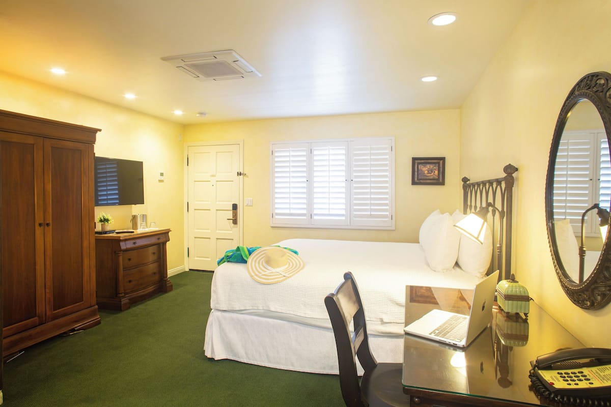 A hotel room with a bed, a desk with a laptop, a telephone, a mirror, a wardrobe, and a window with blinds.