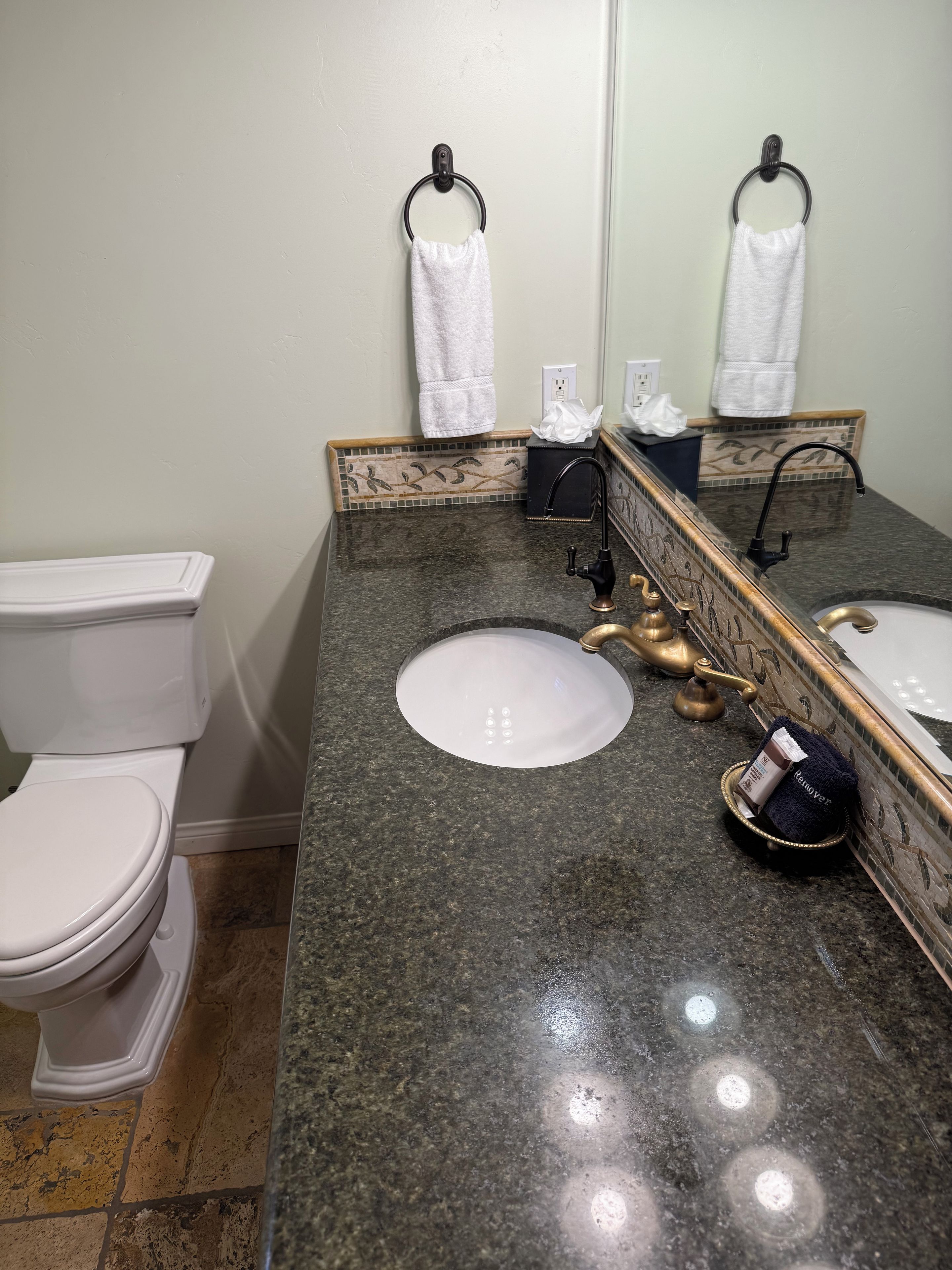 A bathroom with a white toilet, a sink with a gold faucet, and a mirror with a towel hanging on the wall.