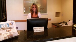 A hotel reception desk with a computer monitor, a newspaper, and a sign stating "NO PETS - NO CHECKS - NO LATE CHECK-OUTS". - Castillo Inn at the Beach (Santa Barbara)