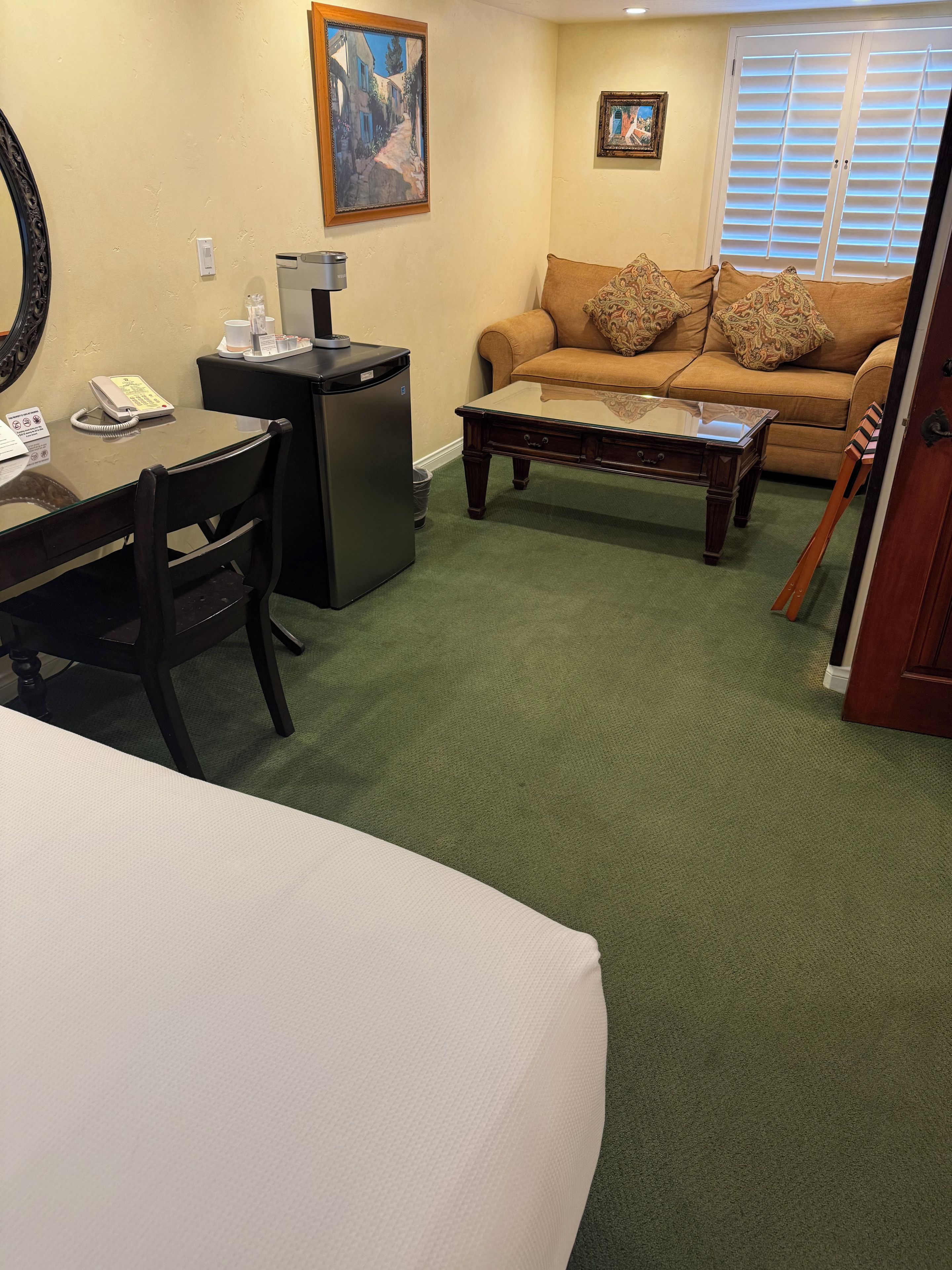 A hotel room with a bed, a desk with a telephone, a refrigerator, a coffee maker, a sofa with patterned cushions, and a painting on the wall.