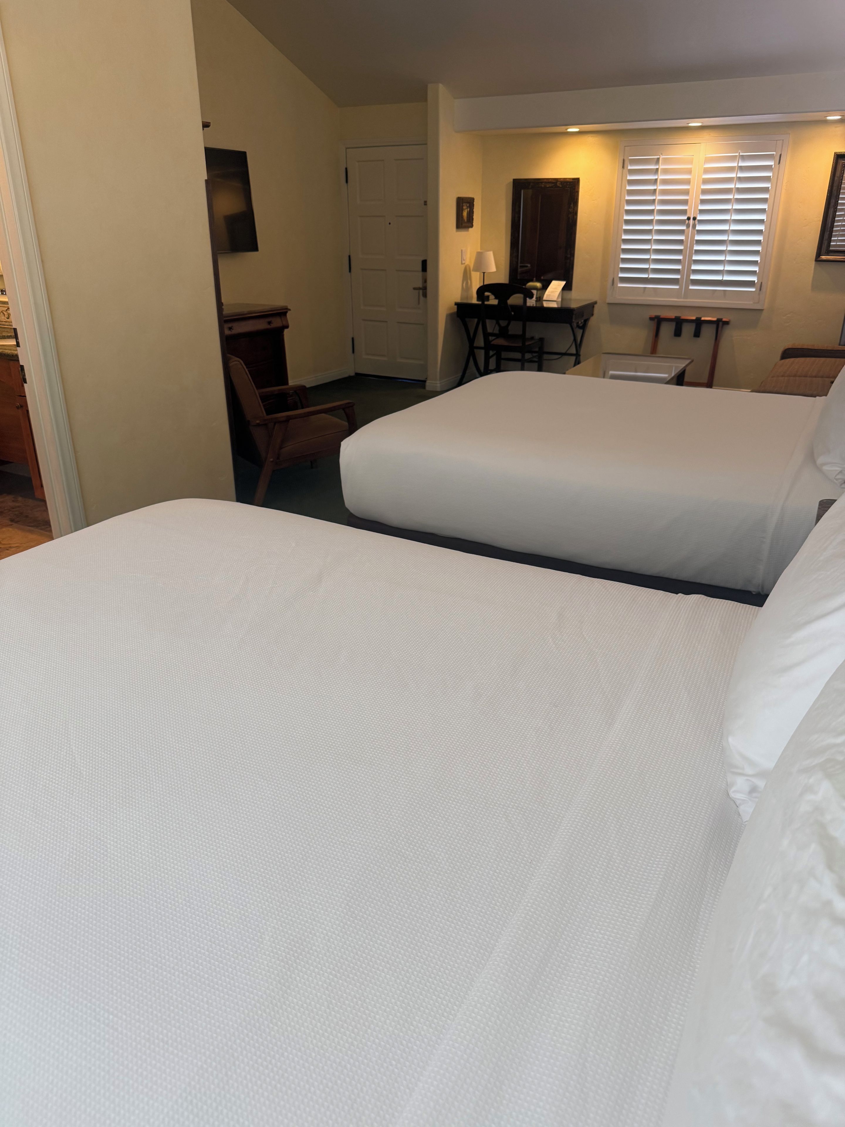 A hotel room with two beds, a desk, a chair, and a window with blinds.