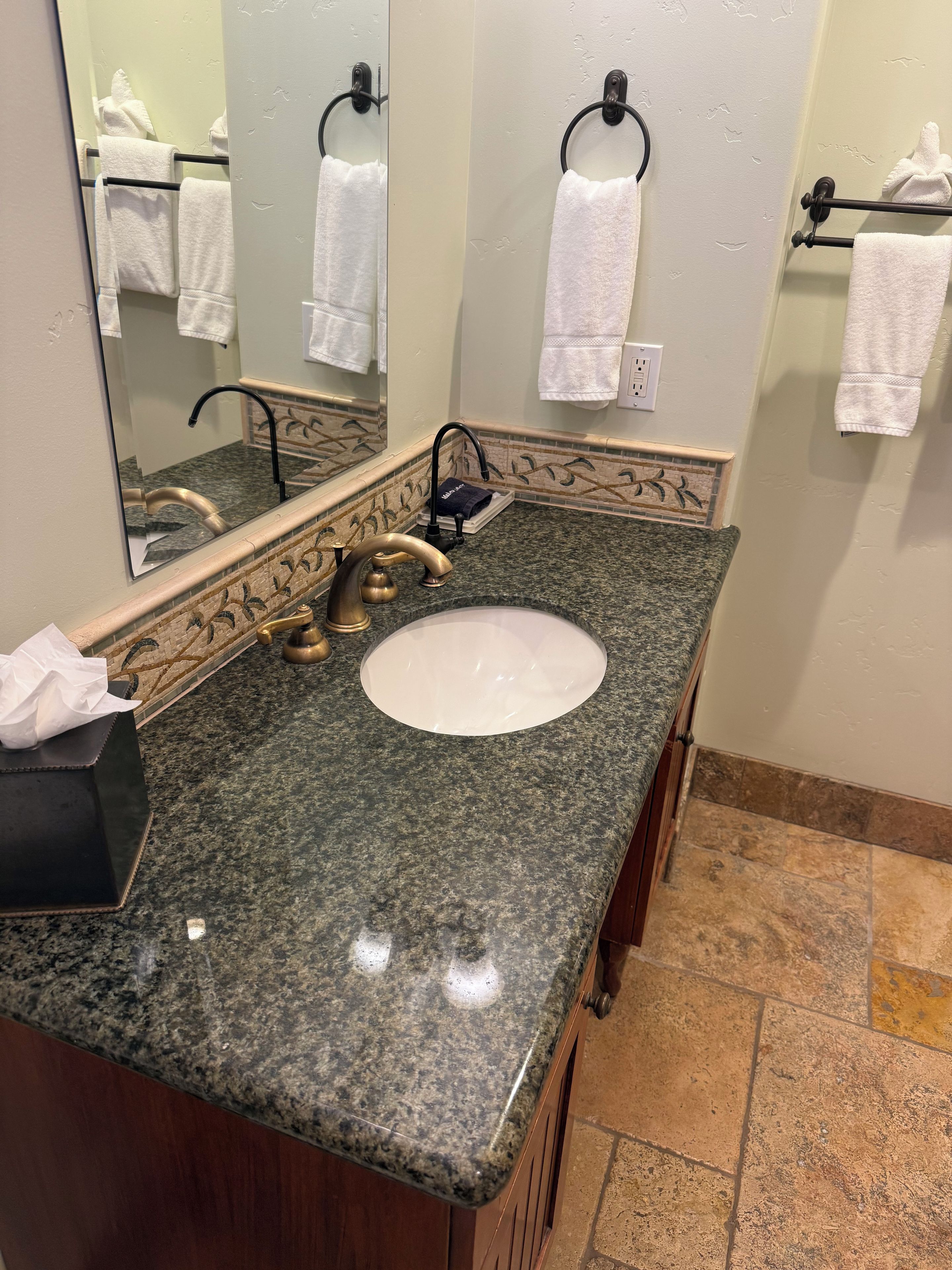 Un baño con encimera de granito, un lavabo blanco, un grifo dorado, un espejo y un perchero para toallas.