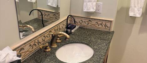 Un baño con encimera de granito, un lavabo blanco, un grifo dorado, un espejo y un perchero para toallas.