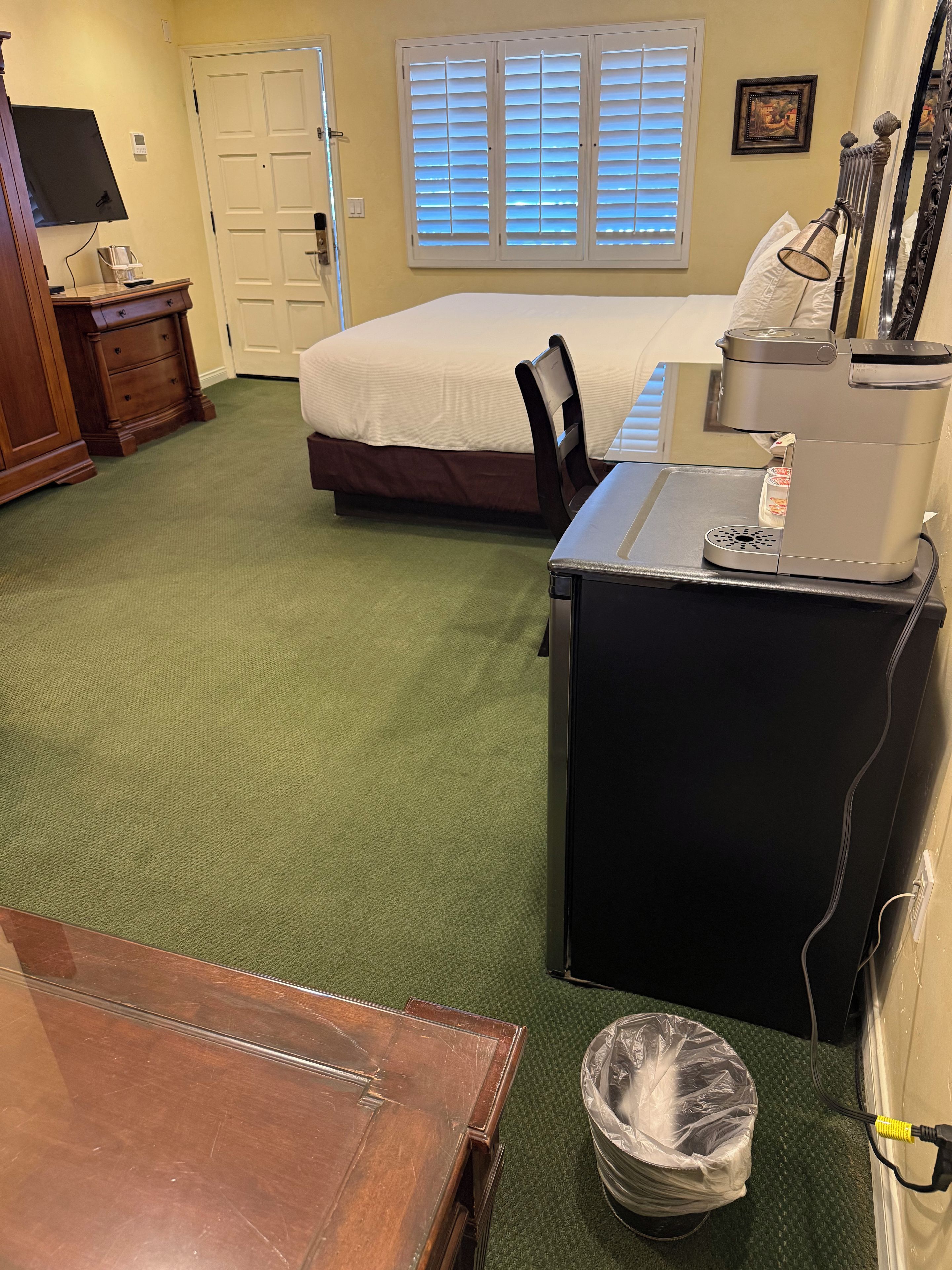 A hotel room with a bed, a desk with a coffee maker, a trash bin, and a TV on a dresser.