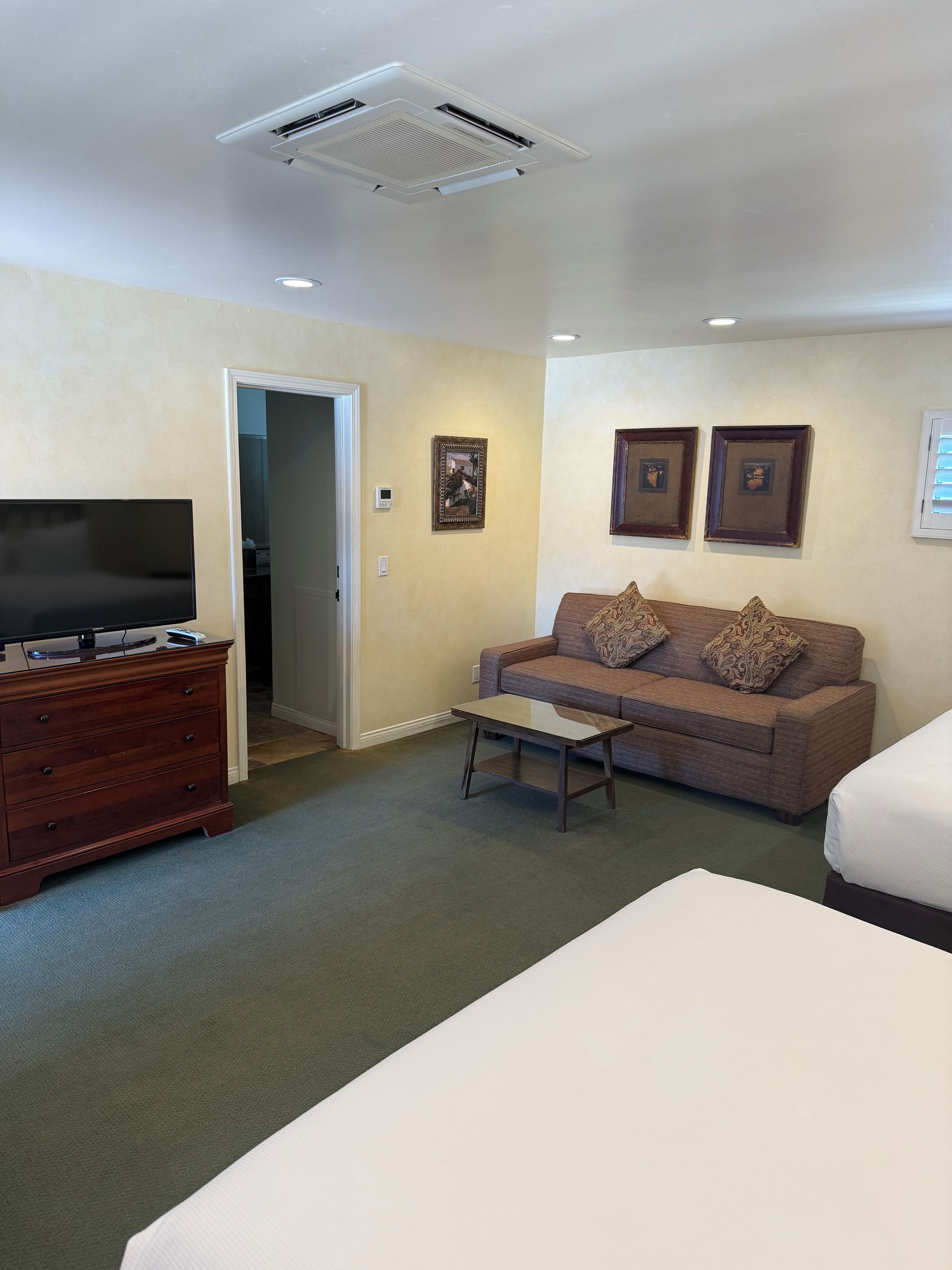 A hotel room with a flat-screen TV, a wooden dresser, a sofa with patterned cushions, a small table, and a bed.