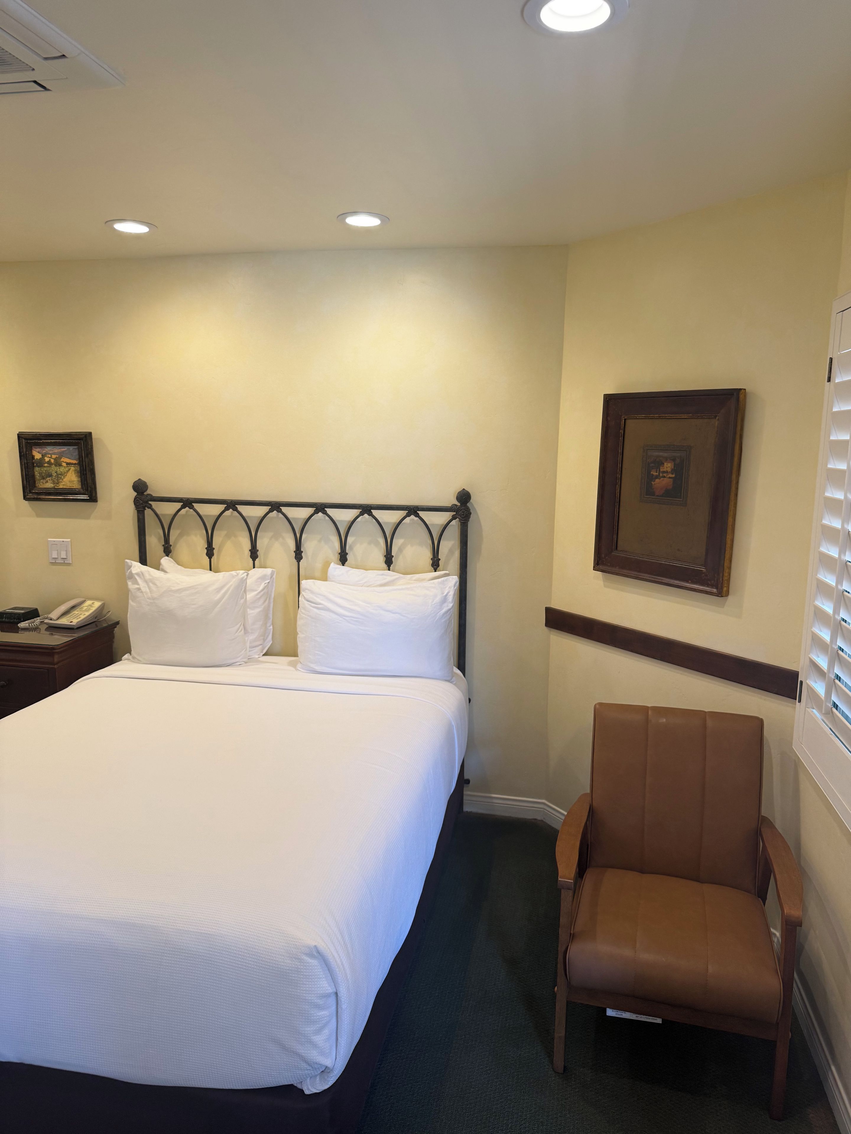 A hotel room with a bed, a chair, a nightstand with a phone, and two wall-mounted pictures.