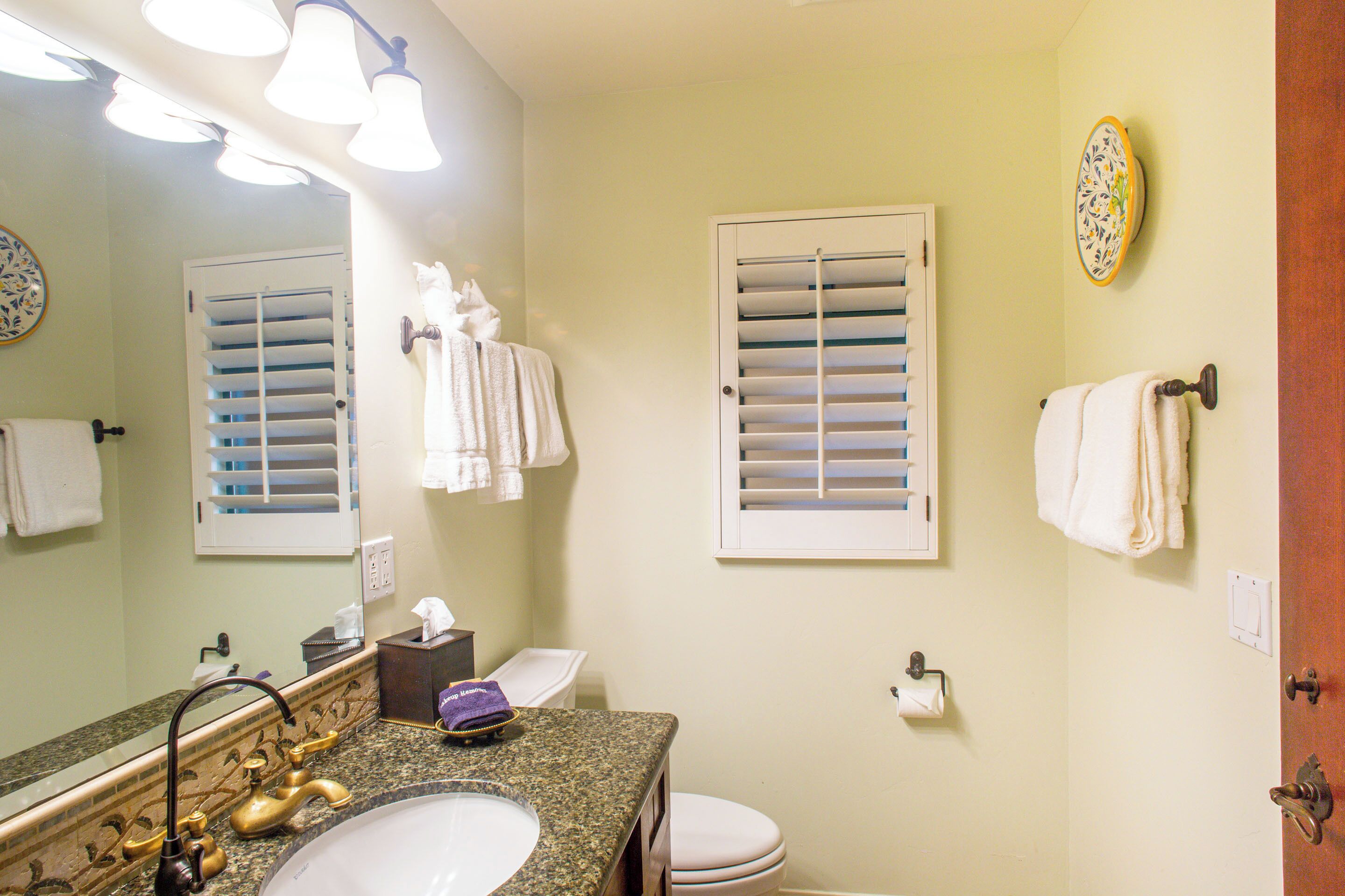 A bathroom with a sink, mirror, towel rack, toilet, and window with blinds.
