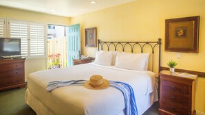 A neatly made bed with a hat and a striped towel, a wooden nightstand with a lamp, and a television on a dresser. - Castillo Inn at the Beach (Santa Barbara)