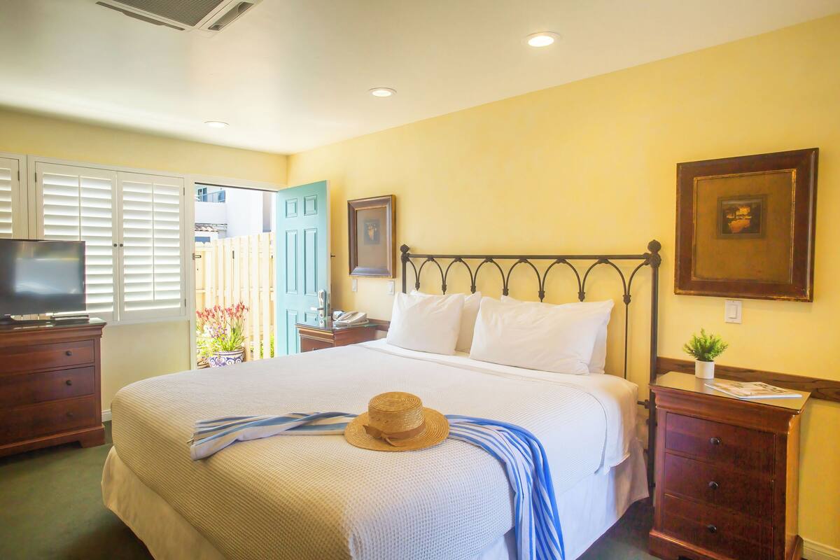 A neatly made bed with a hat and a striped towel, a wooden nightstand with a lamp, and a television on a dresser.