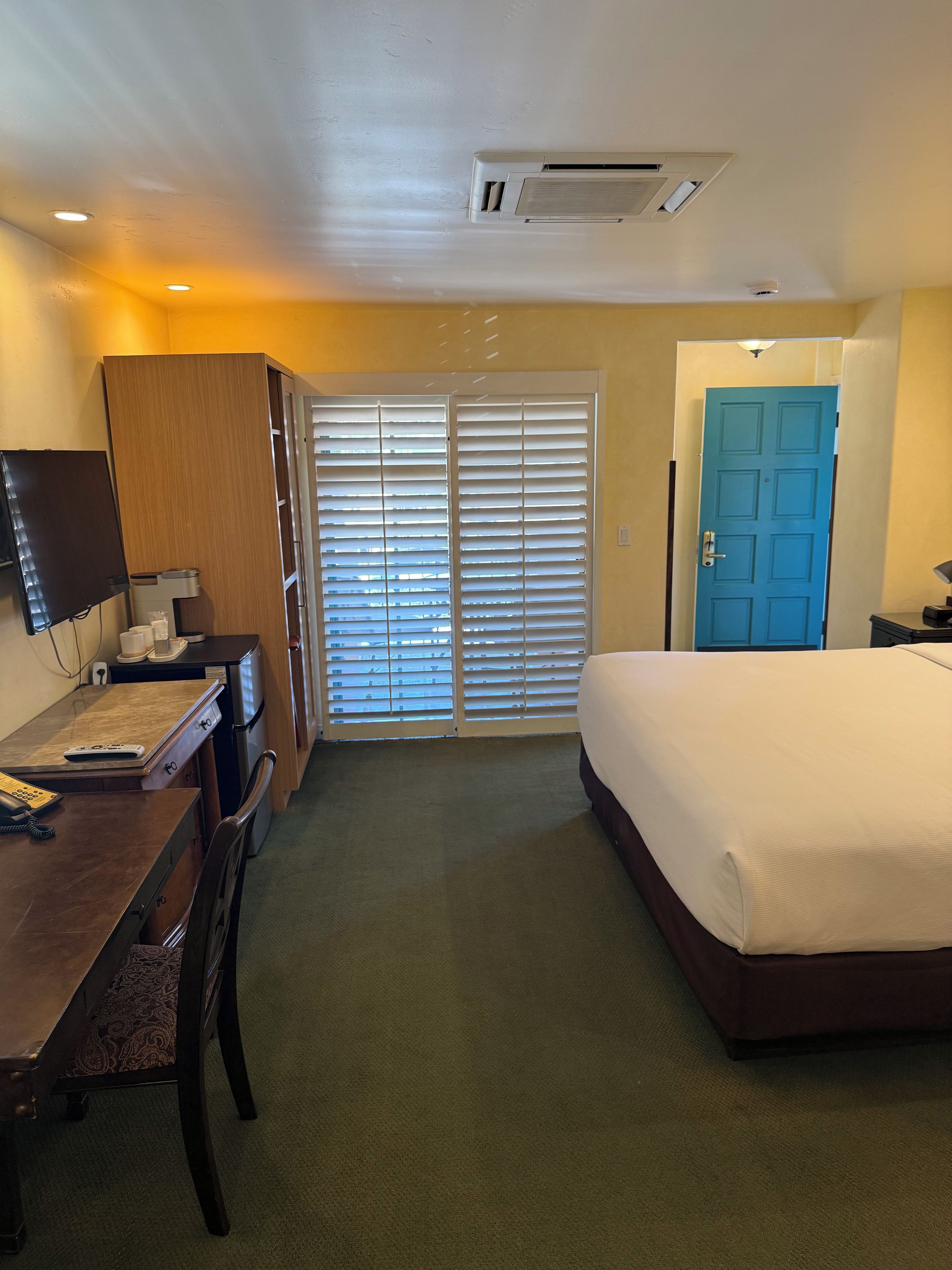 A hotel room with a bed, desk, television, and a window with blinds.