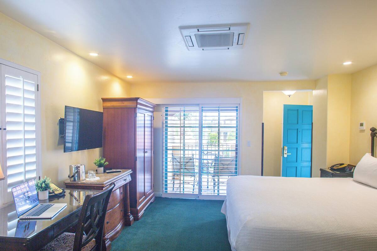 A hotel room with a bed, a desk with a laptop, a TV, and a window with blinds.