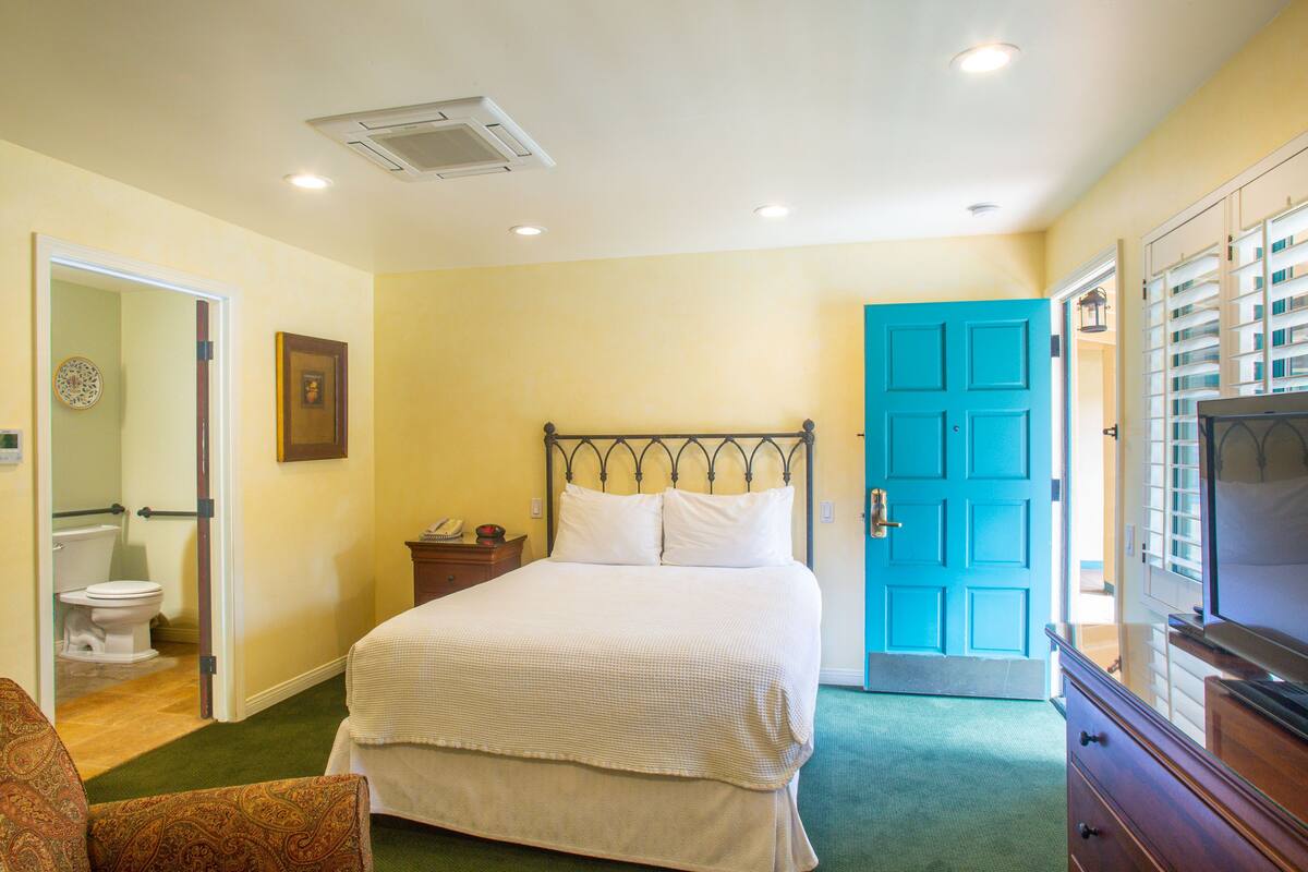 A bedroom with a bed, a television, a dresser, and a bathroom visible through an open door.