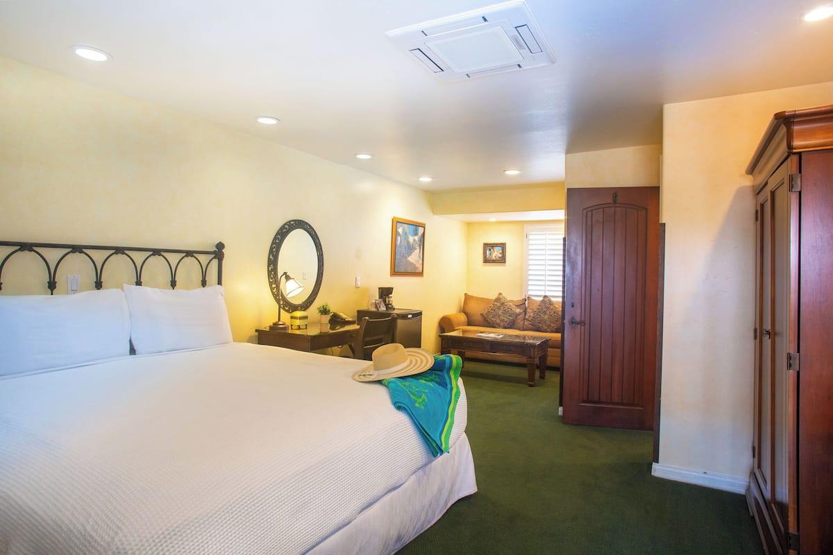 A hotel room with a large bed, a desk with a chair, a sofa, a wardrobe, and a door.