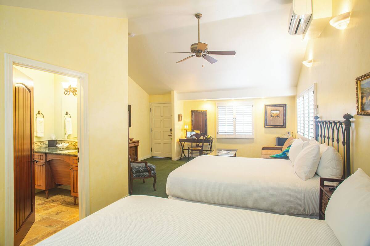 A hotel room with two beds, a desk, a chair, a ceiling fan, and a bathroom with a mirror and towel rack.