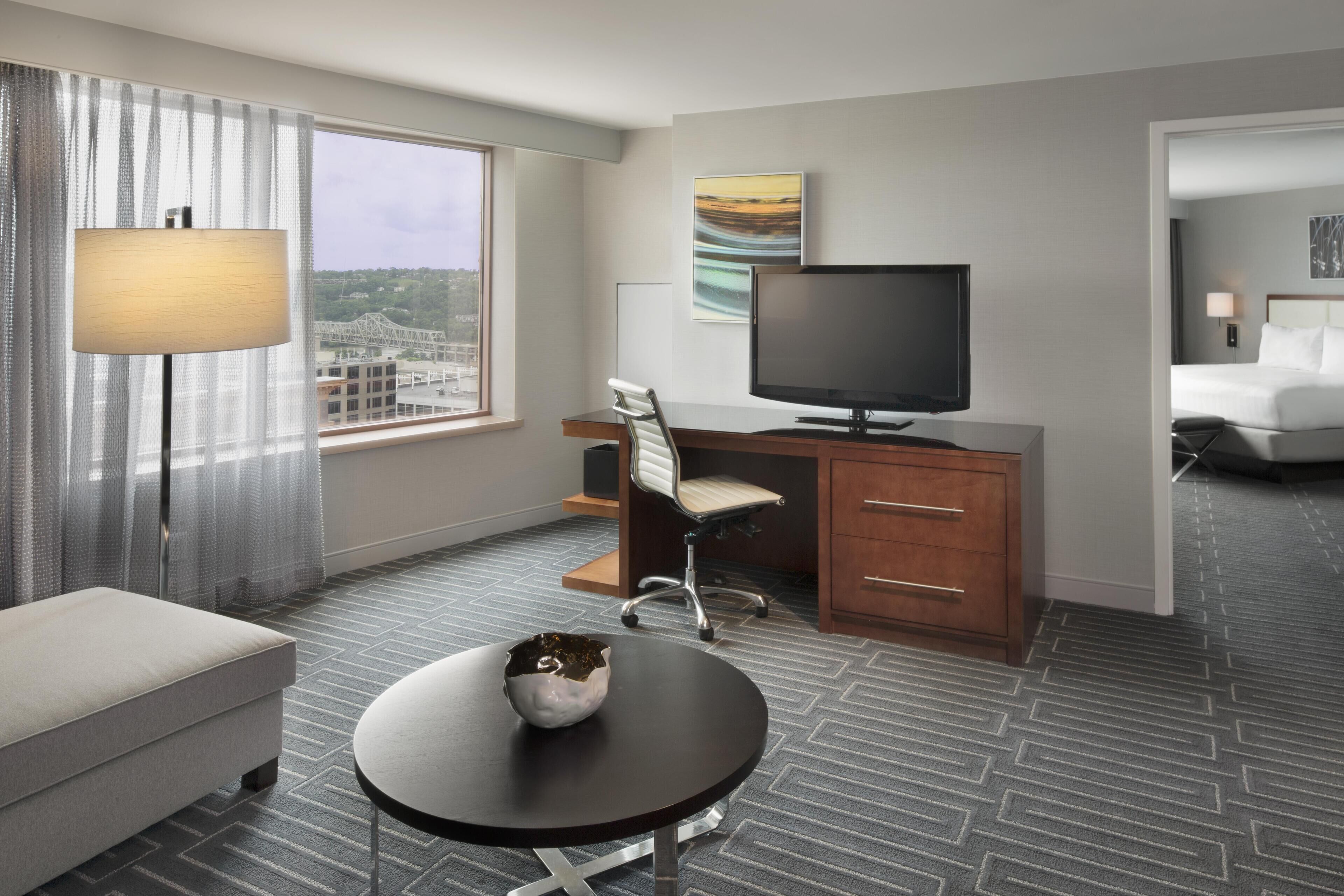 A hotel room with a bed, a desk with a TV, a chair, a small table with a decorative item, and a view of a cityscape through the window.