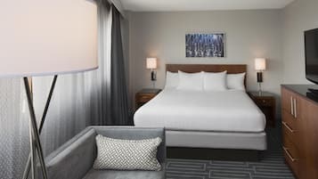 Ett hotellrum med en stor säng, två sängbord med lampor, en soffa med ett mönstrat kuddöverdrag, en tv på ett skåp och en tavla på väggen.
