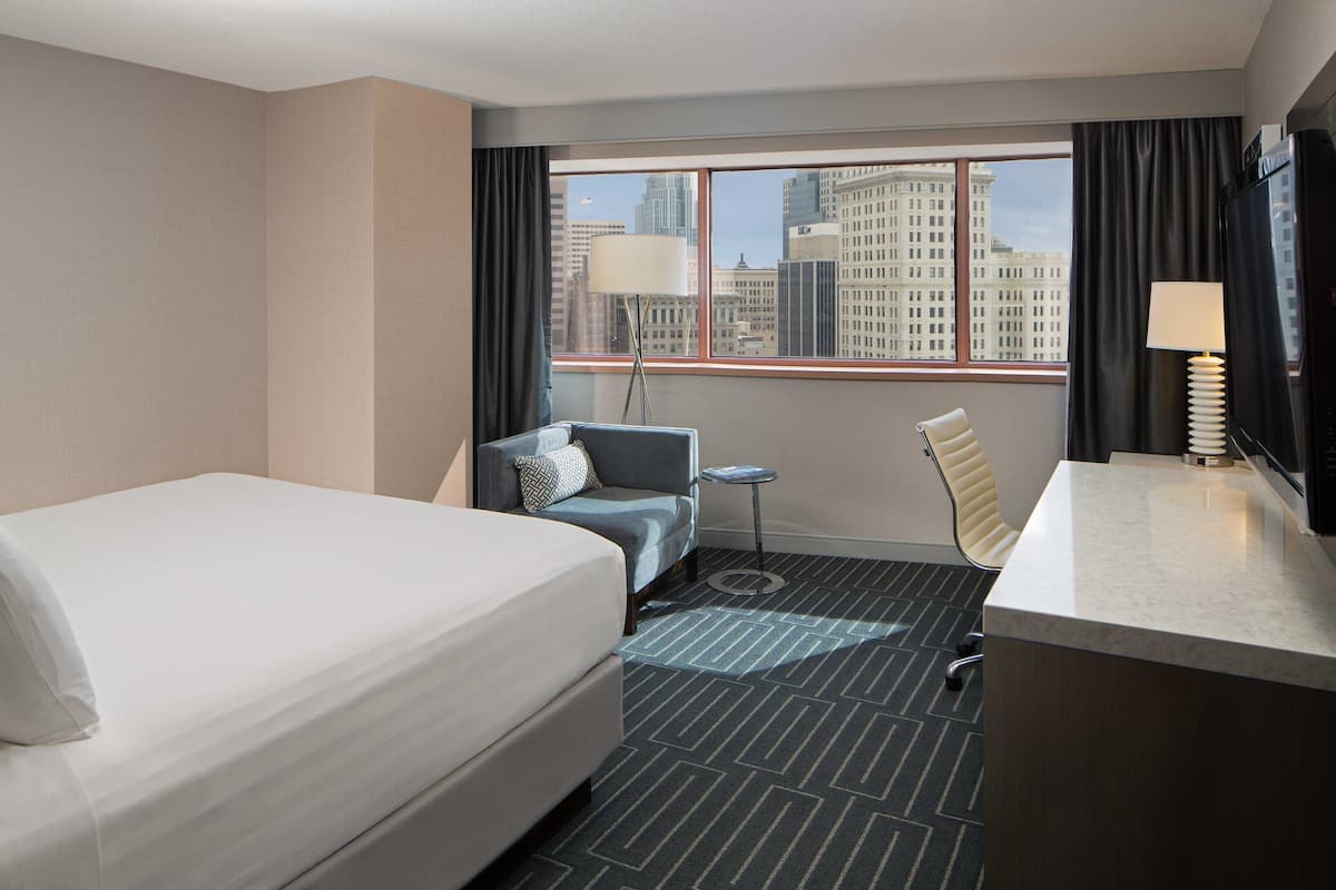 A hotel room with a bed, a desk, a chair, a small table, and a view of a cityscape through large windows.