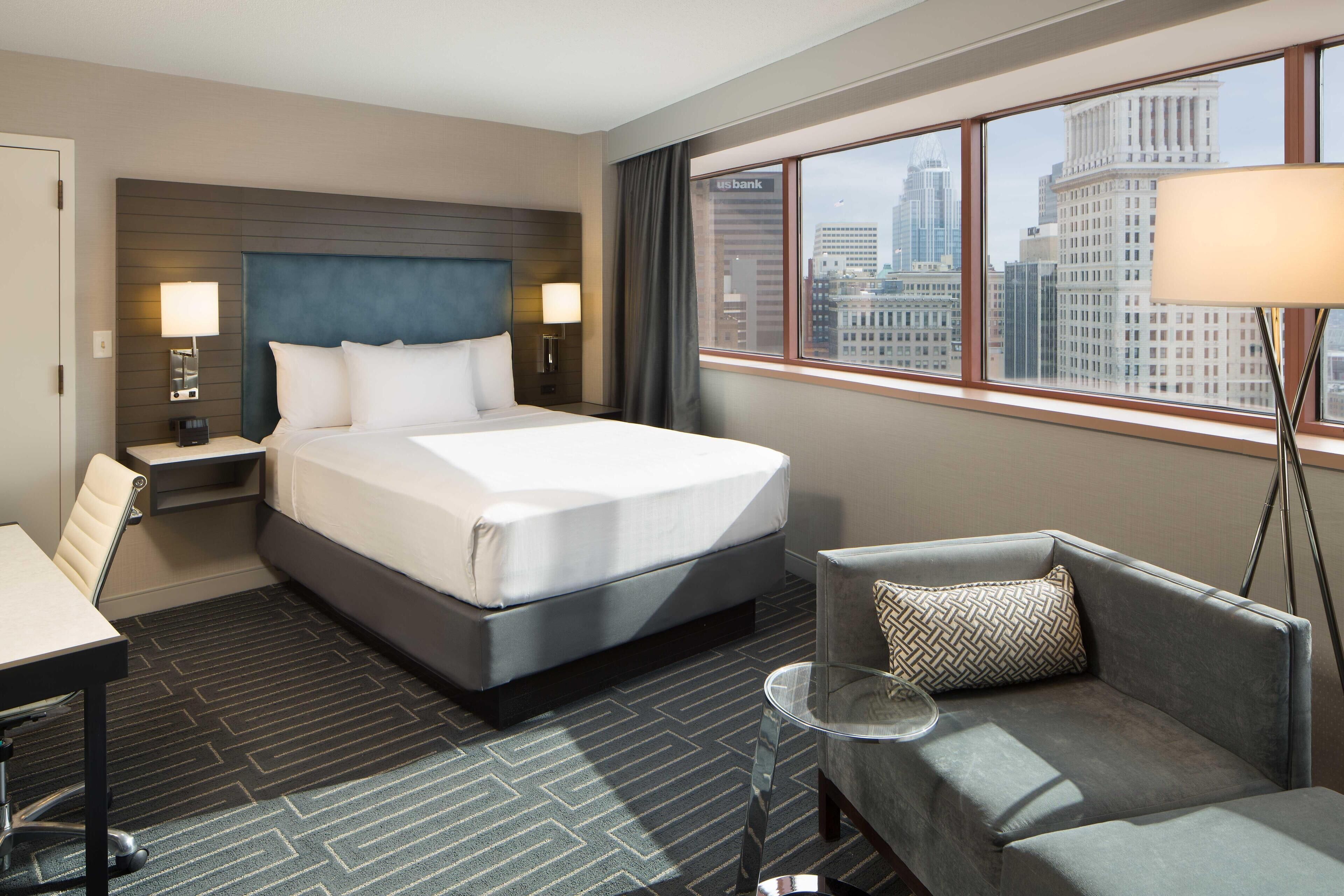 A hotel room with a large bed, a desk, a chair, and a view of the city skyline through the window.