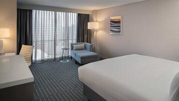 A hotel room with a bed, a desk, a chair, a small table, a lamp, and a window with curtains.