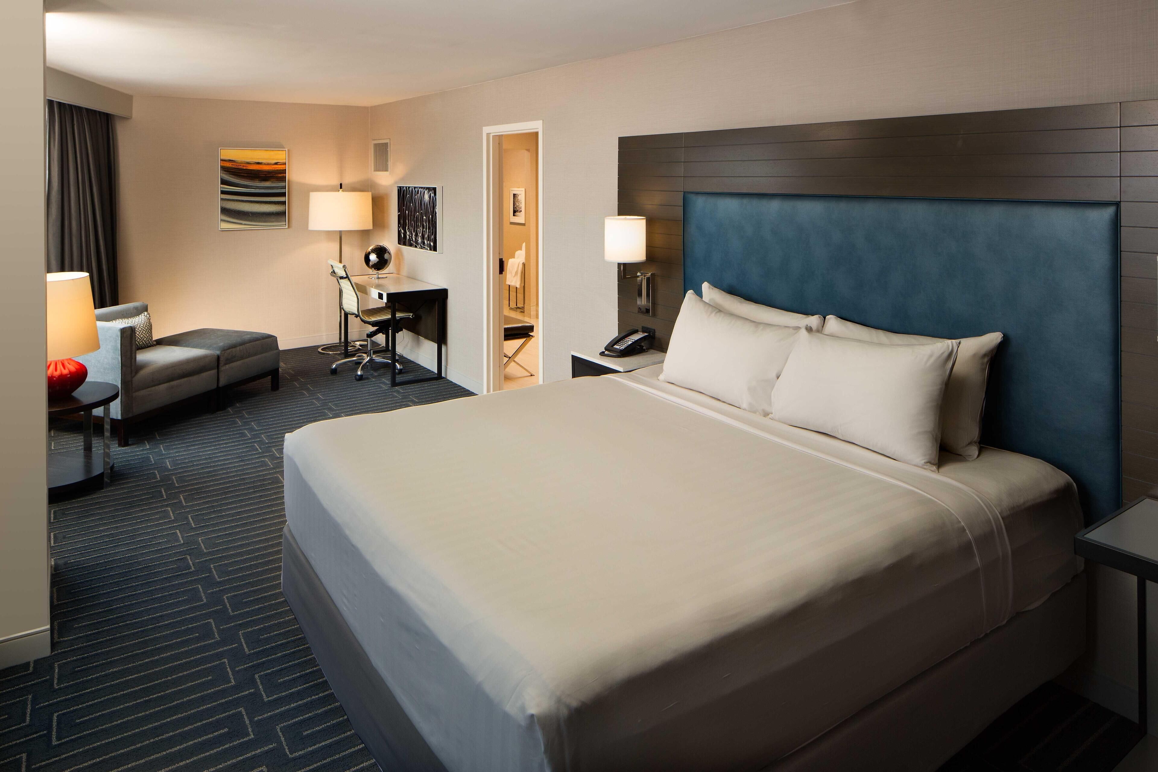 A hotel room with a large bed, a desk with a chair, a small table, and a lamp.