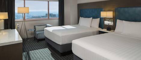 A hotel room with two beds, a desk, a chair, a small table, and a view of the city.