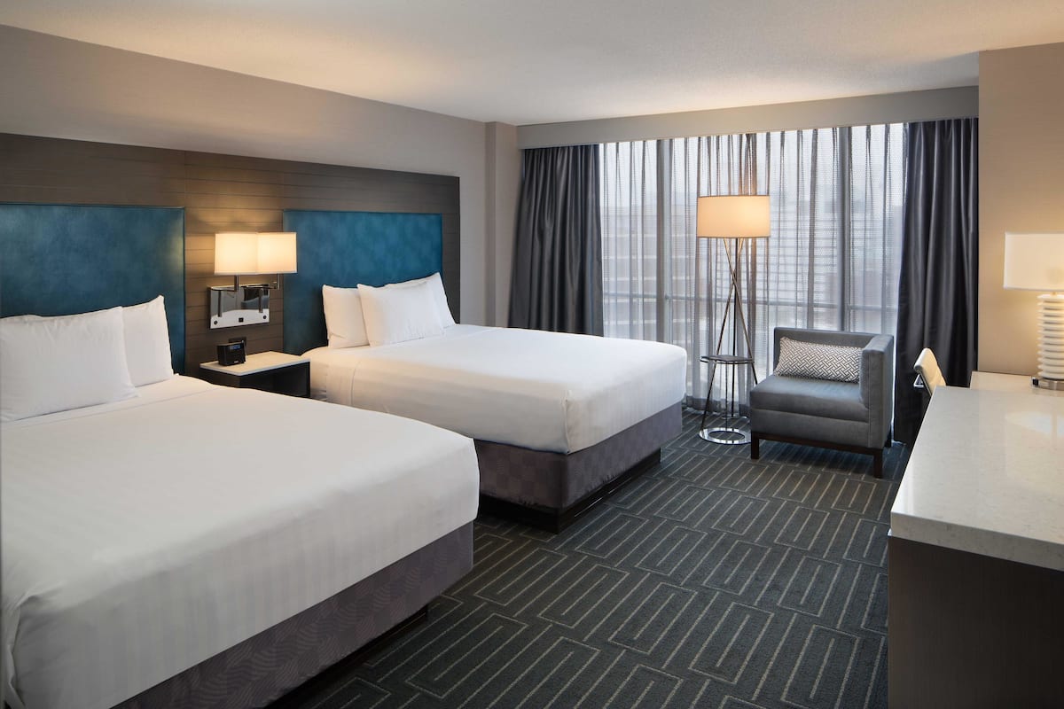 A hotel room with two beds, a desk, a chair, a lamp, and a large window with curtains.
