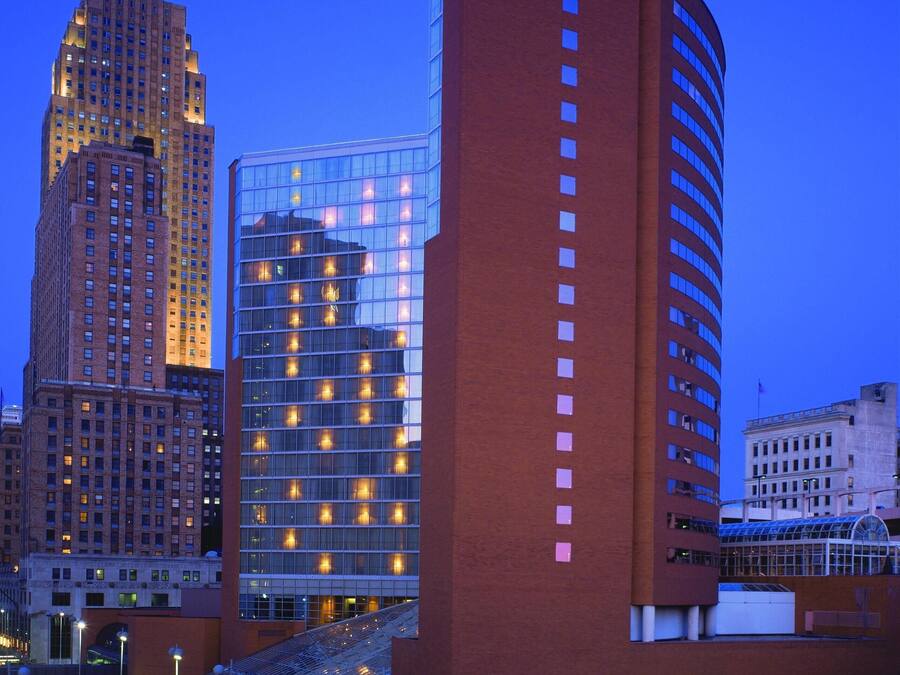 Um hotel com uma placa 'HYATT' em destaque, fachada de vidro moderna e um edifício de tijolos clássico nas proximidades.