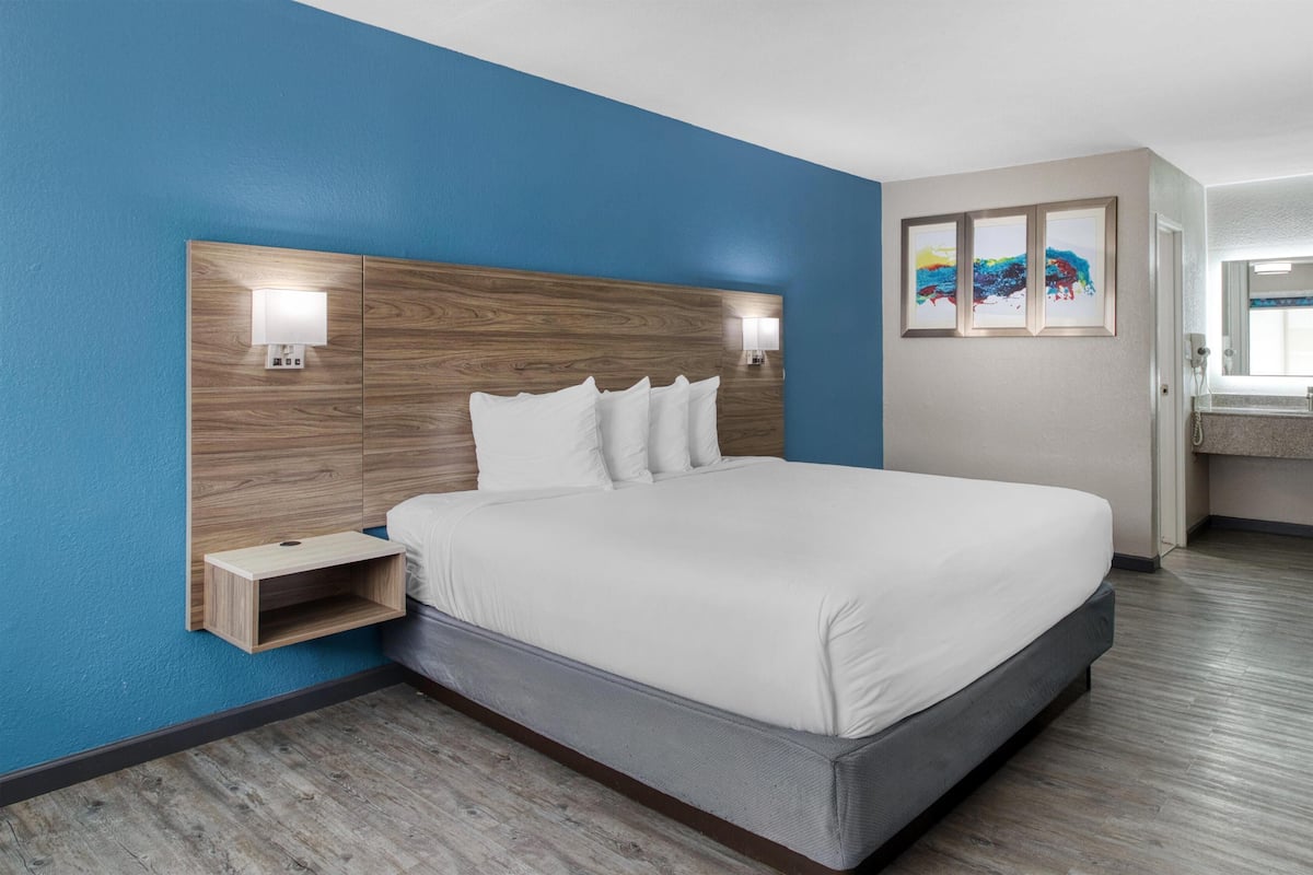 A modern hotel room with a large bed, a wooden headboard, a bedside table with a lamp, and a small shelf.