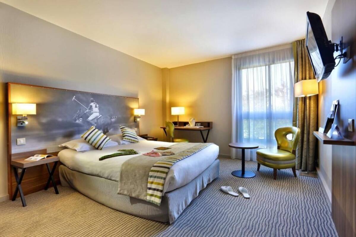 A hotel room with a bed, bedside tables, a desk, a chair, a TV, and a large window with curtains.