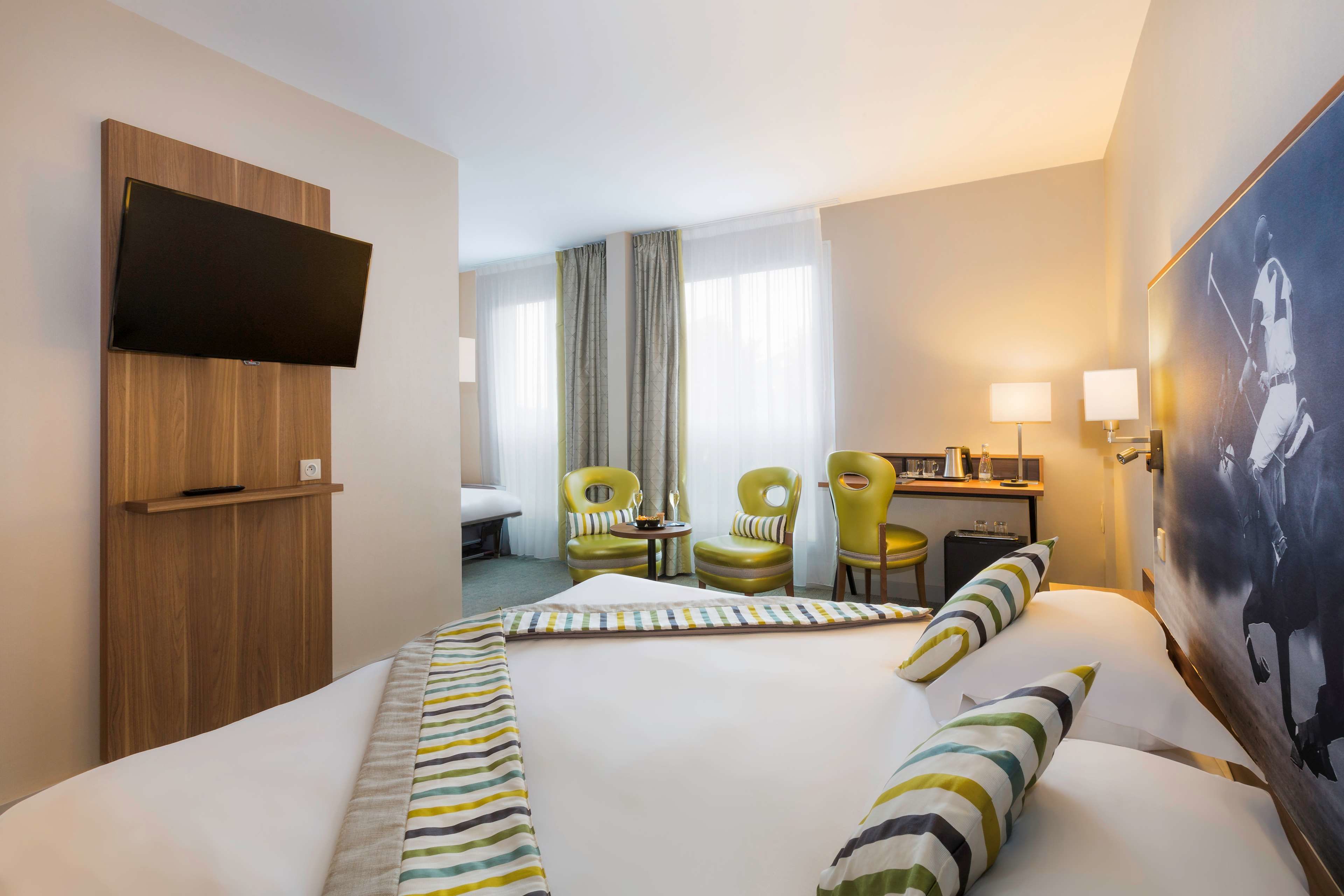A hotel room with a large bed, two green chairs, a desk with a lamp, and a TV mounted on the wall.