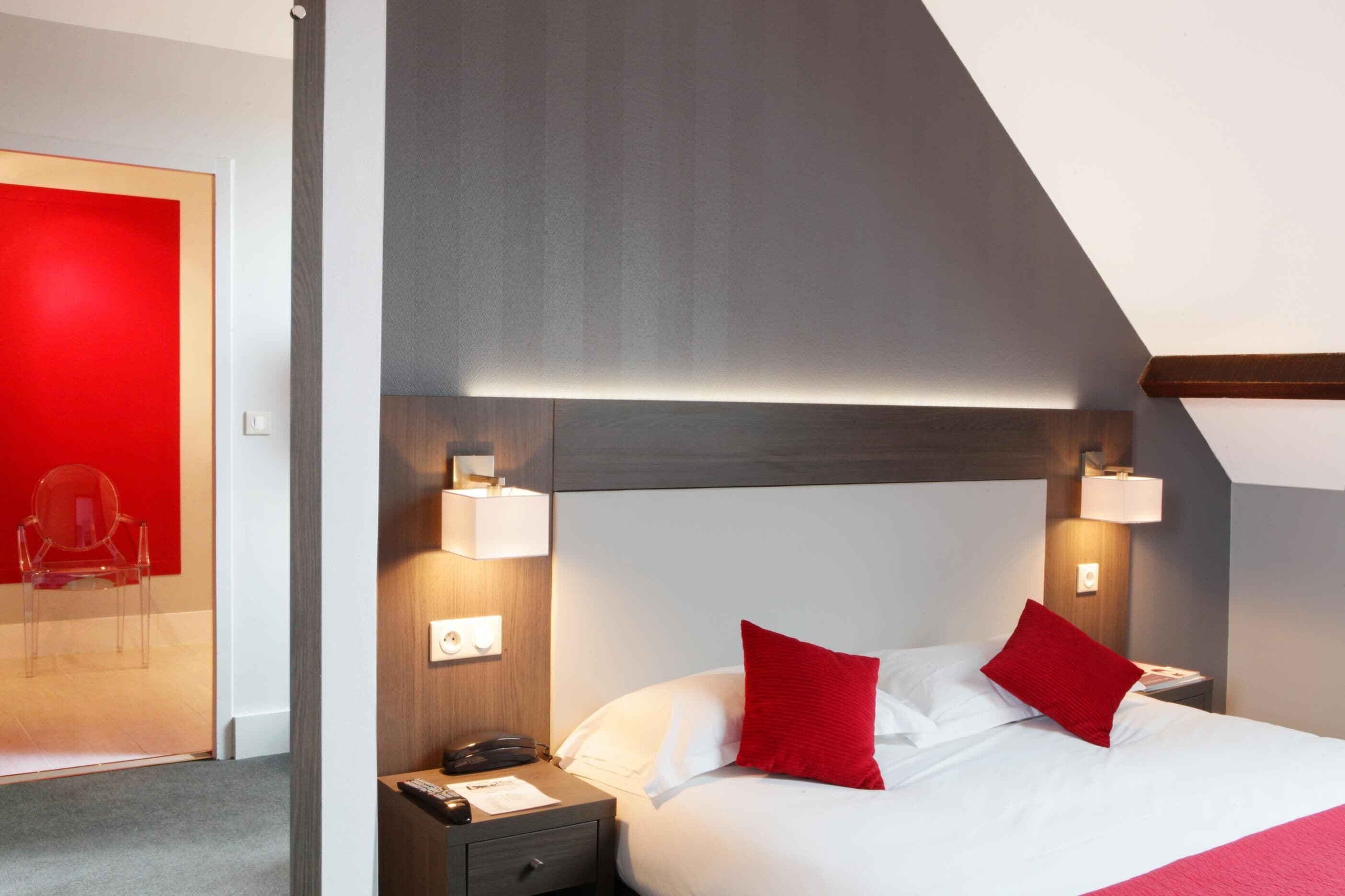 A hotel room with a modern design, featuring a bed with red pillows, a nightstand with a lamp, and a wooden headboard.