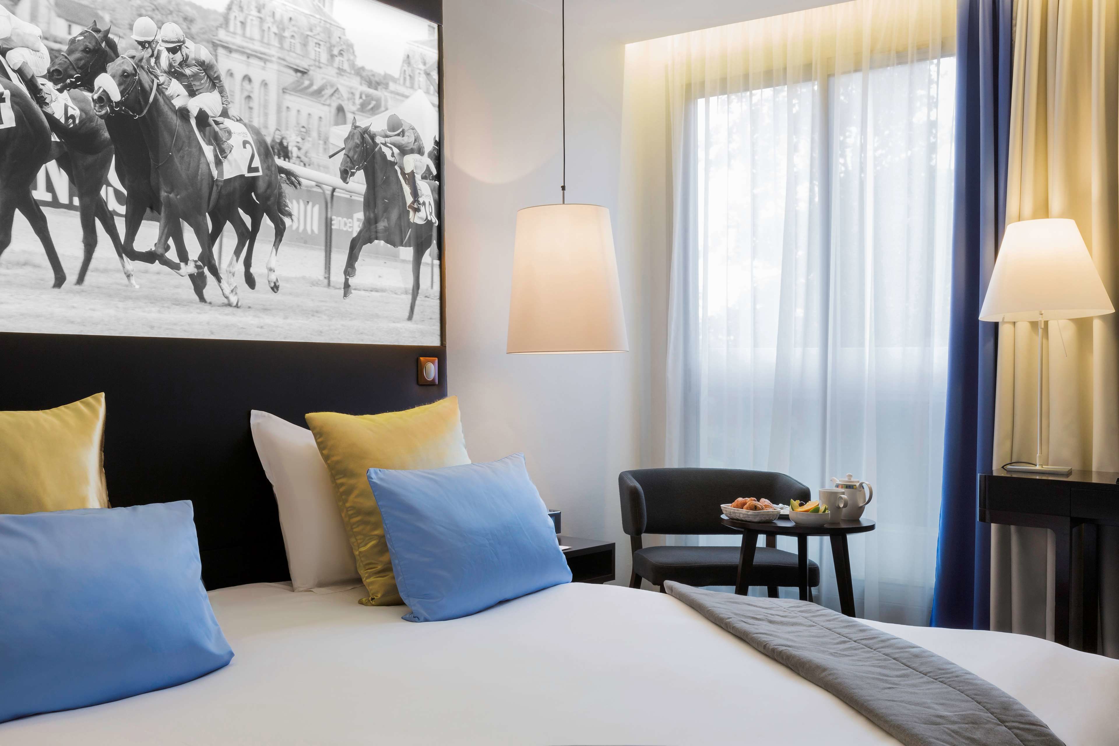 Quarto de hotel com uma cama grande, uma escrivaninha com cadeira, uma mesinha com uma tigela de frutas e uma luminária na parede.