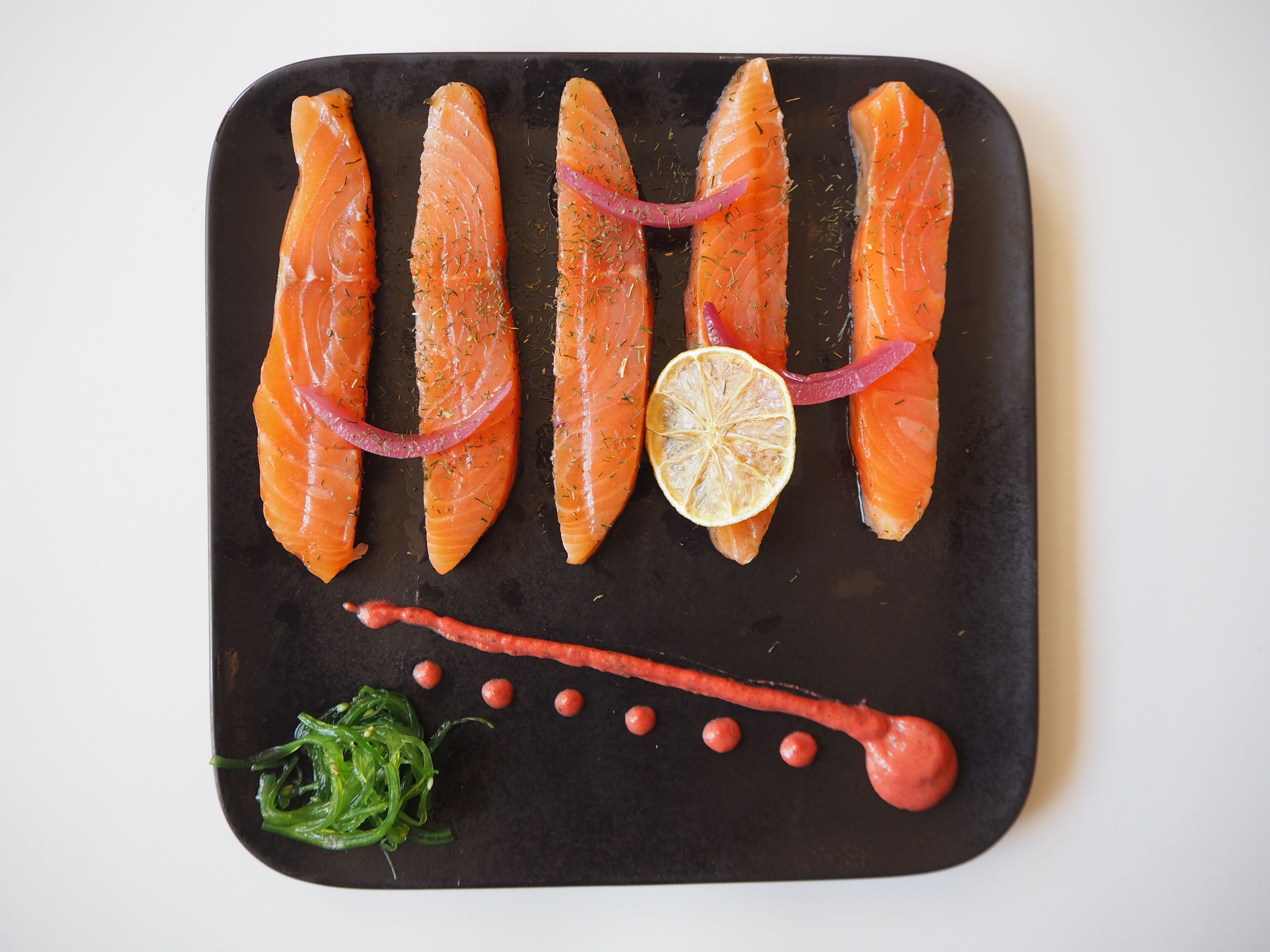 A black rectangular plate with five pieces of salmon, garnished with dill and red onion slices, and accompanied by a lemon slice and a red sauce with small red dots.