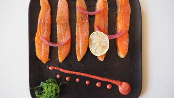 A black rectangular plate with five pieces of salmon, garnished with dill and red onion slices, and accompanied by a lemon slice and a red sauce with small red dots.