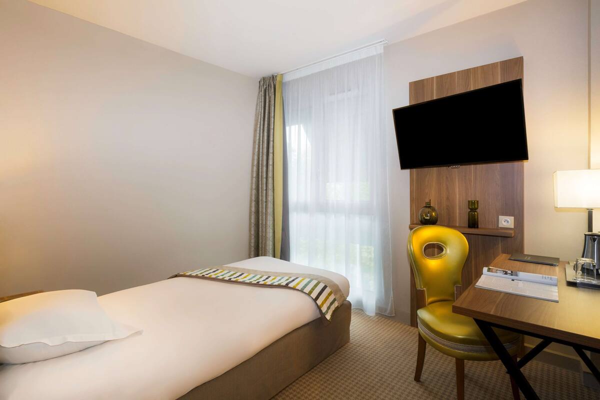 A hotel room with a bed, a desk, a chair, a flat-screen TV, and a window with curtains.