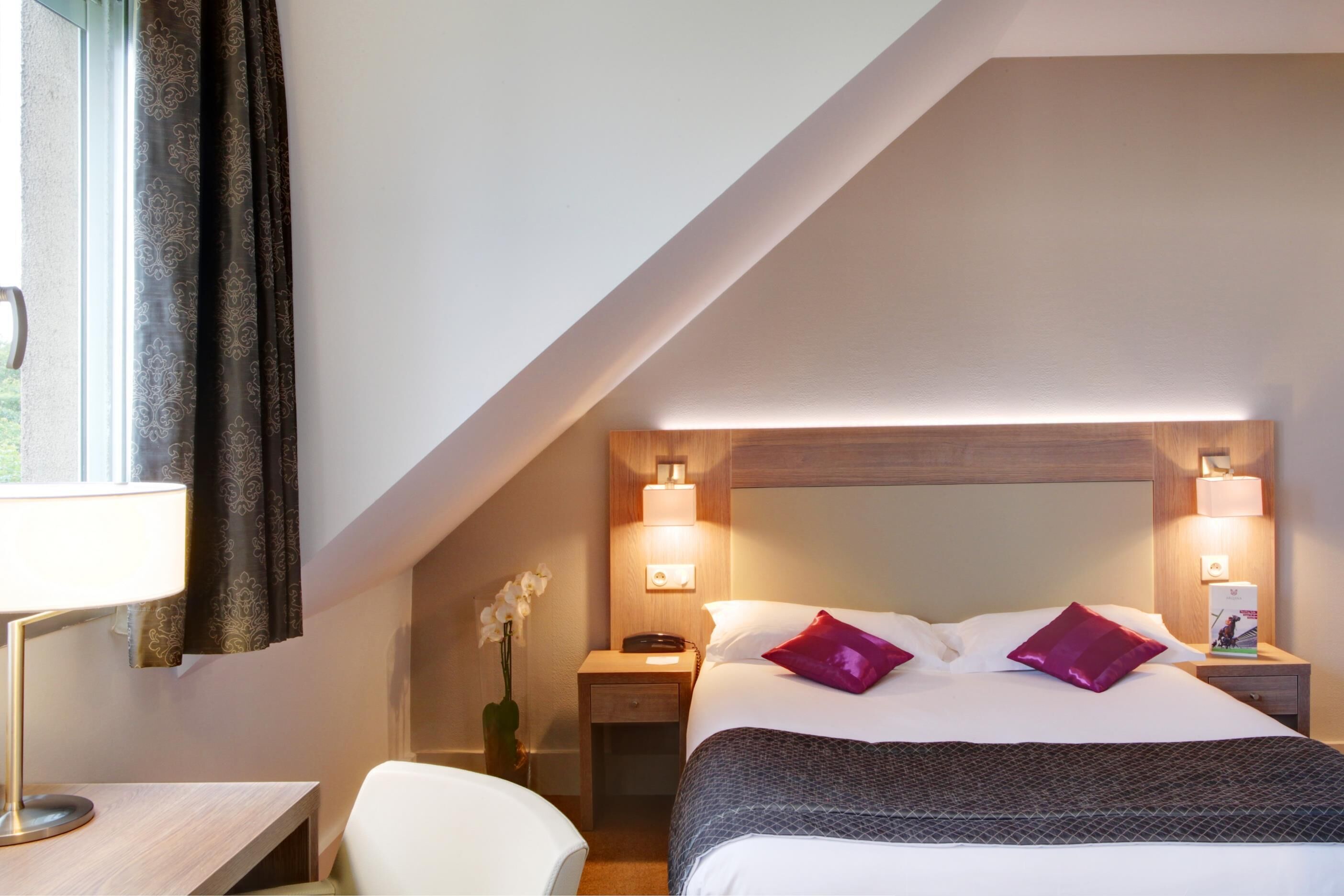A hotel room with a sloped ceiling, a bed with white and purple bedding, two bedside lamps, a nightstand with a phone and a small table, and a chair.